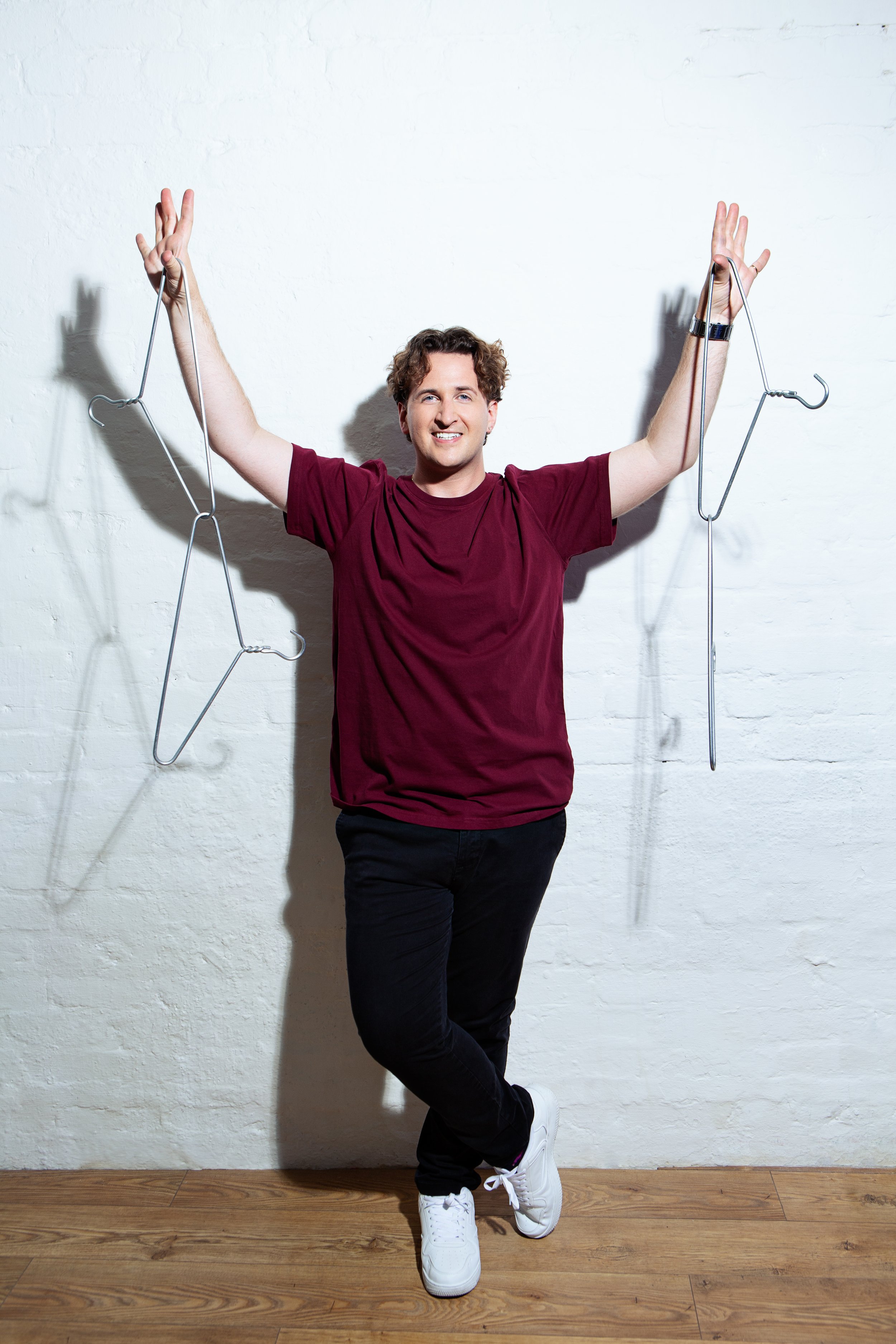 A young man standing against a white brick wall, wearing a burgundy T-shirt, black pants, and white sneakers, holding wire coat hangers with his arms raised.