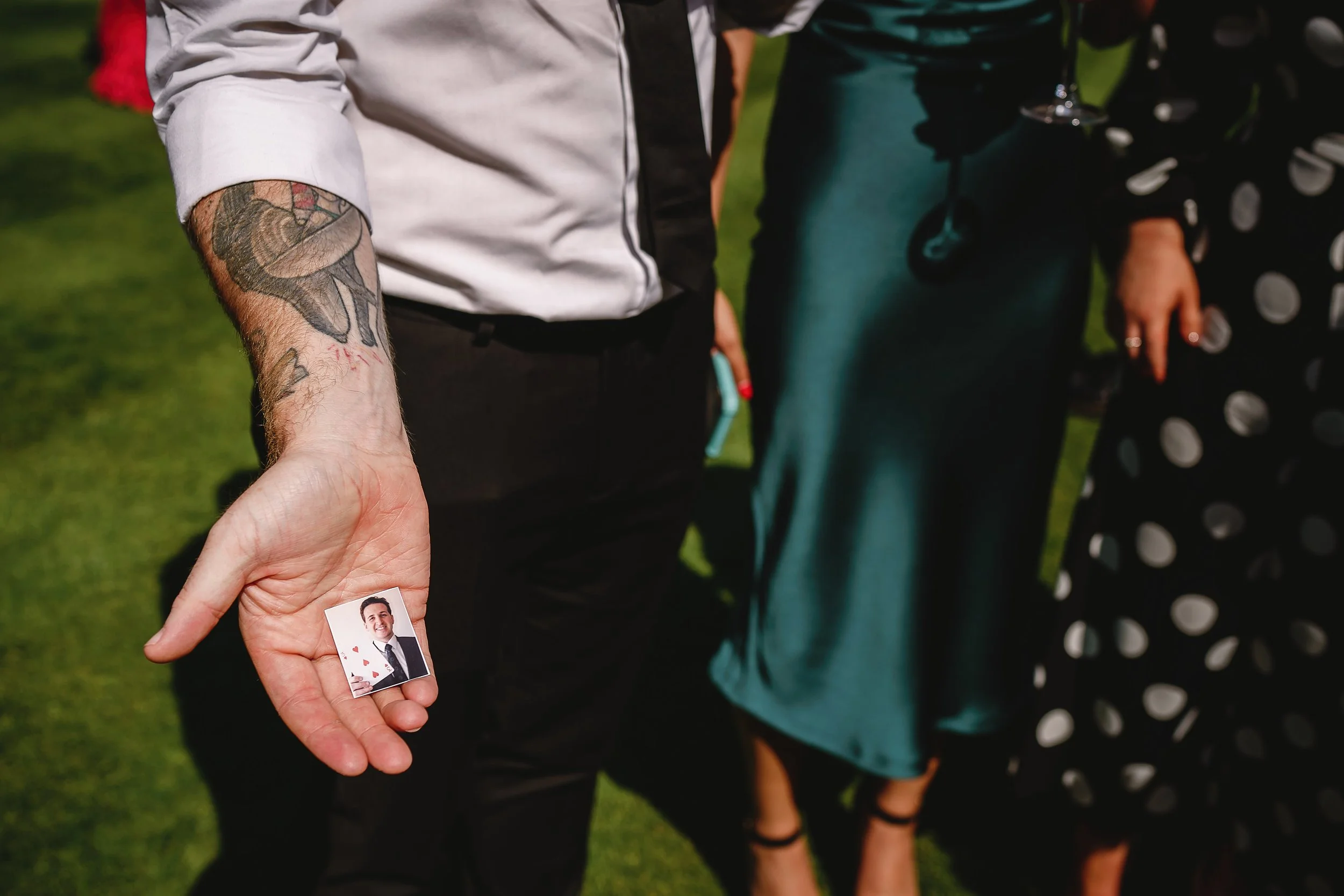 Close-up of a person's hand holding a small photo of a smiling young man in a suit, with a lizard tattoo on their arm and people dressed formally in the background.