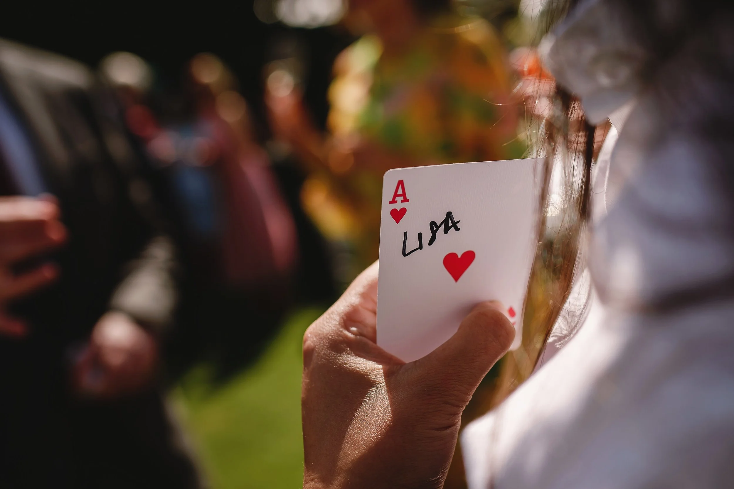 Close-up of a person holding an ace of hearts playing card with the name 'LISA' written on it, in an outdoor setting with blurred people and greenery in the background.
