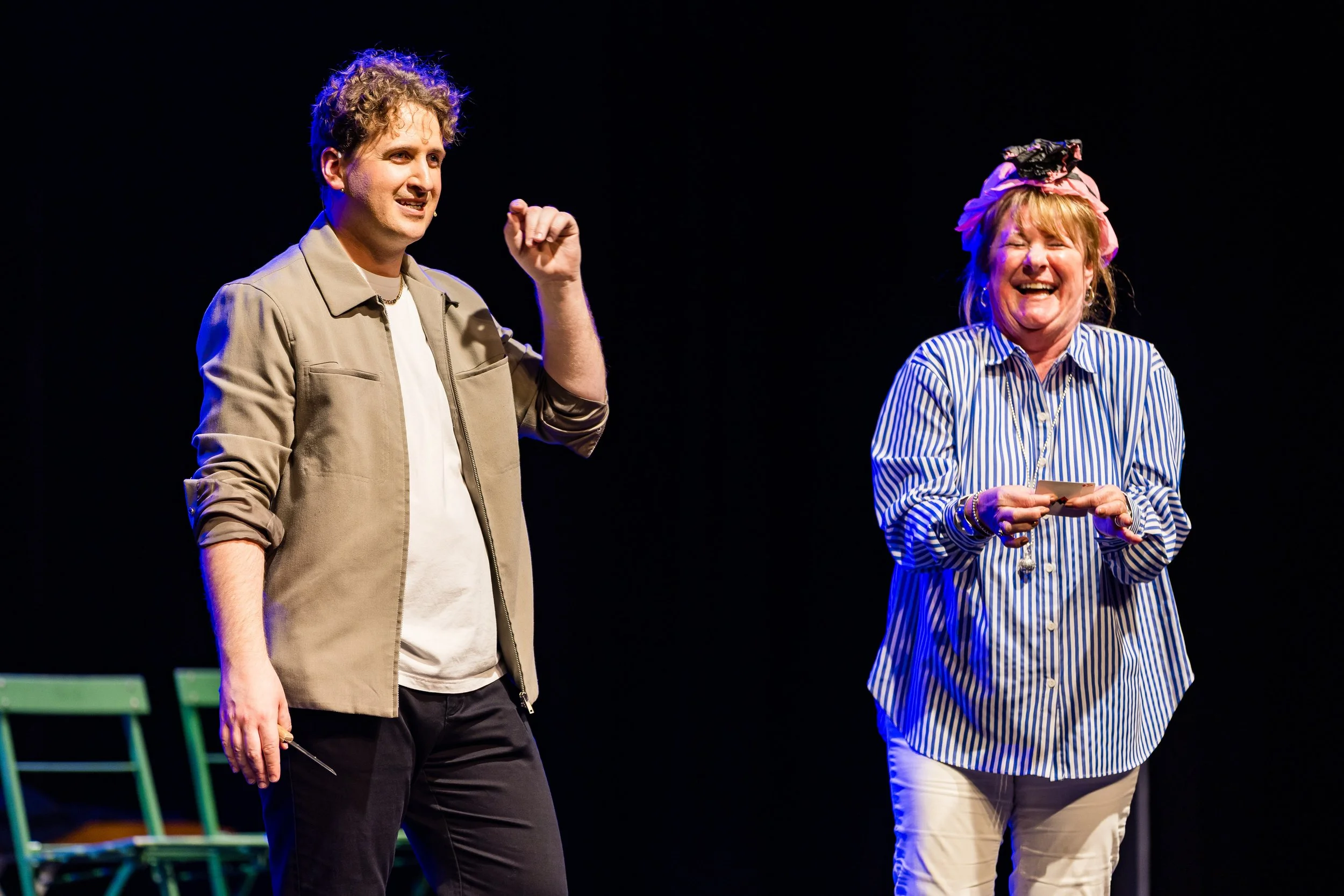 Two people, a man on the left and a woman on the right, are standing on a stage. The man is gesturing with his right hand, wearing a tan jacket and white shirt. The woman is smiling, holding a piece of paper, and wearing a striped blue and white shir