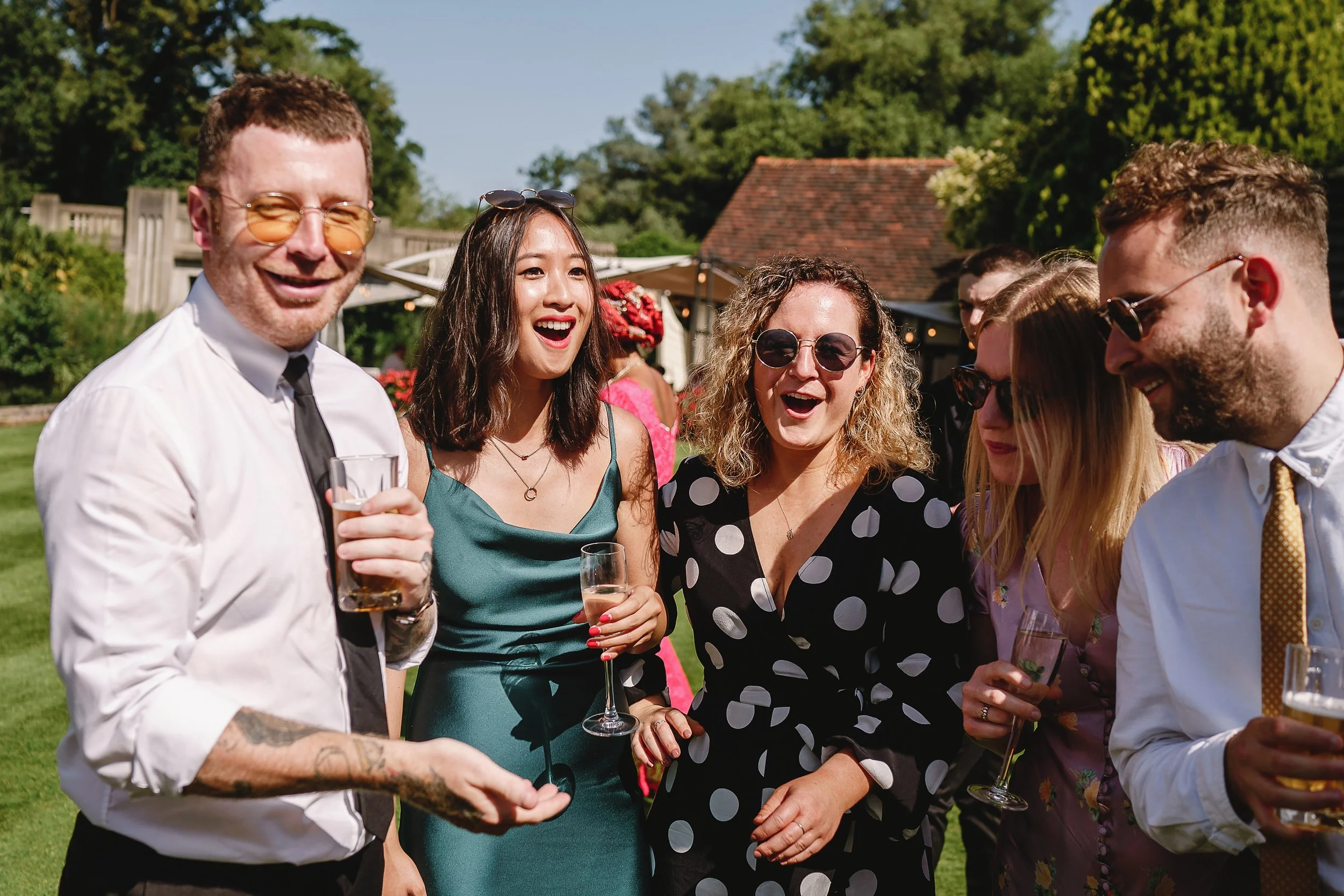Group of people at an outdoor party, smiling and holding drinks, with a sunny backyard and trees in the background.