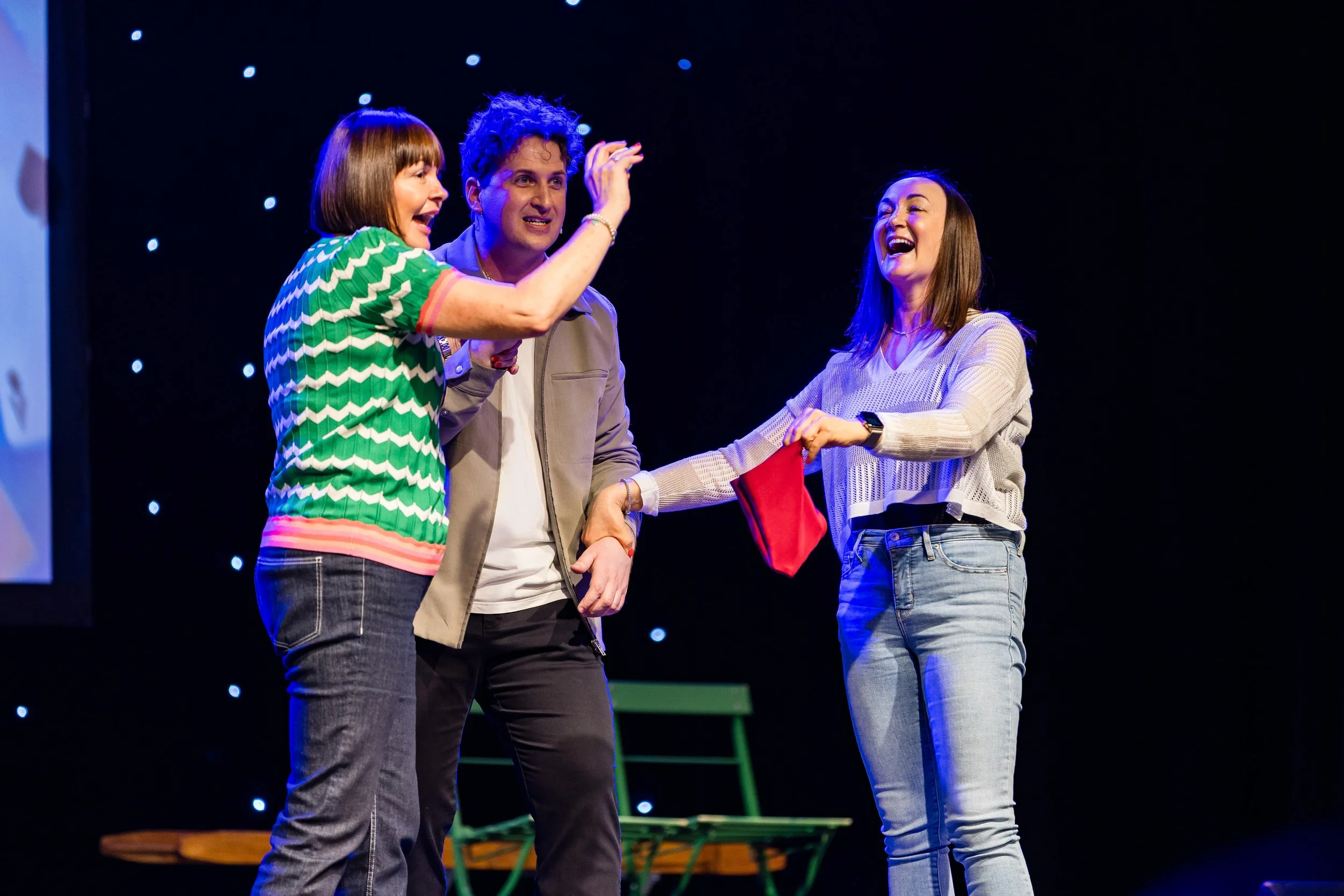 Three women on stage during a presentation or event, one woman is handing a red gift bag to another, with both women smiling and laughing, the third woman looks on with a smile.