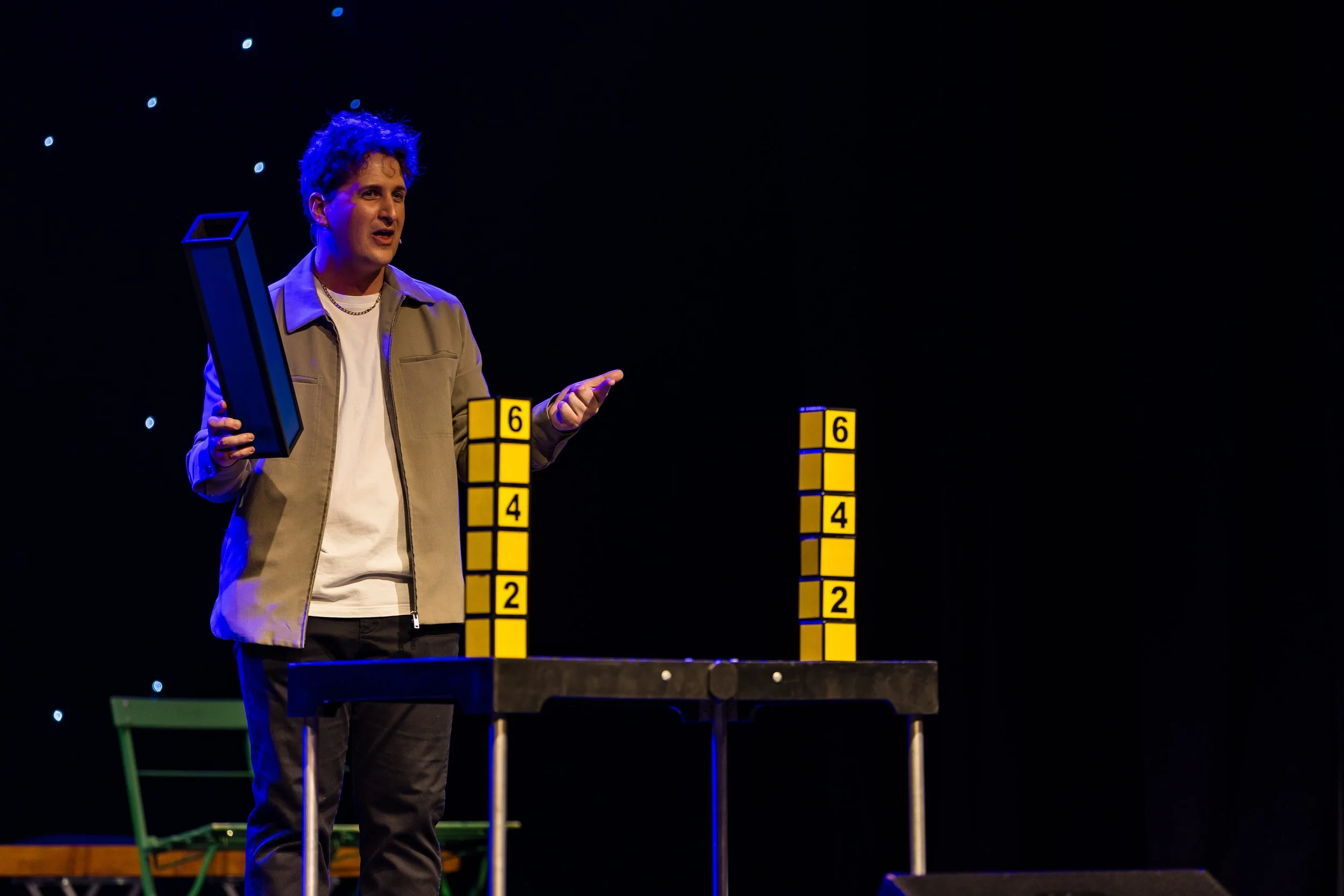 A man with curly hair standing on stage, holding a tall yellow tower block with the number 6. There are two other similar towers with the numbers 6, 4, and 2 visible on a table, with a dark background and stage lights.