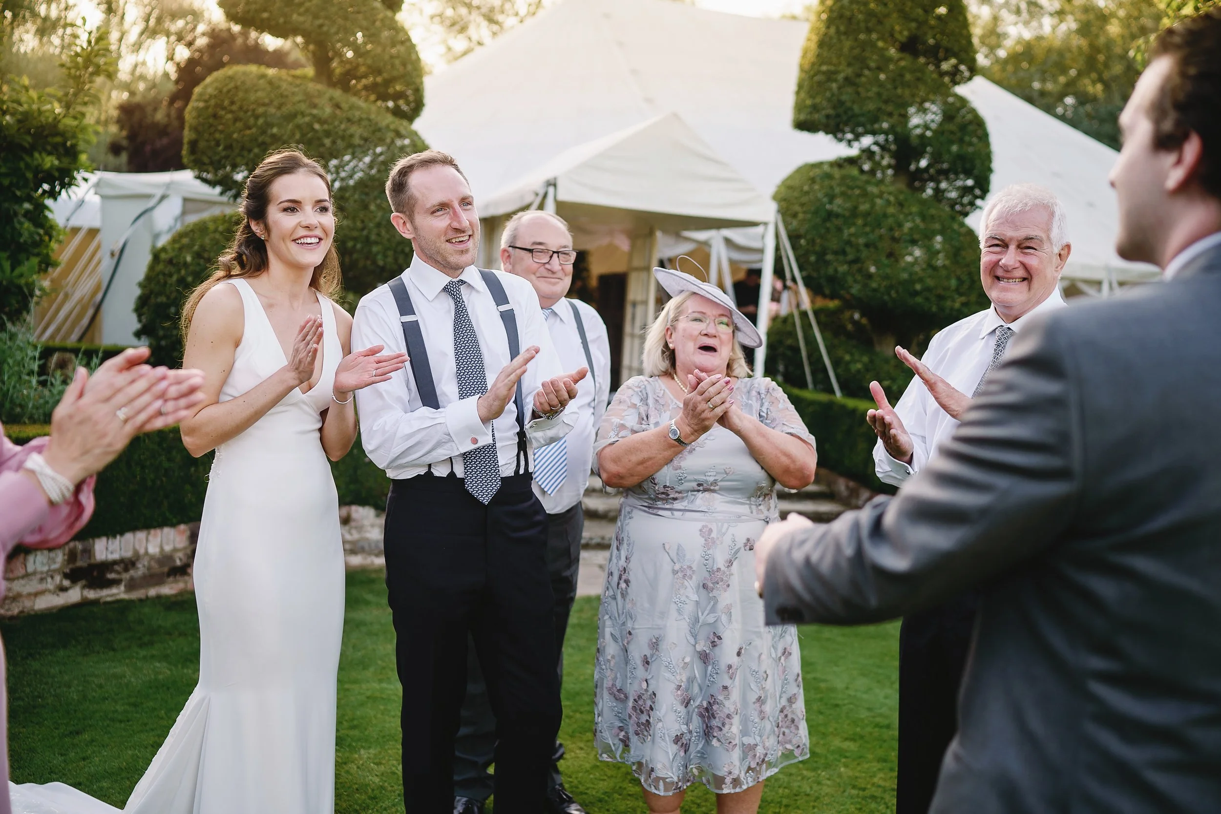 Group of people celebrating at an outdoor wedding or event, smiling and clapping, with a white tent and green trees in the background.