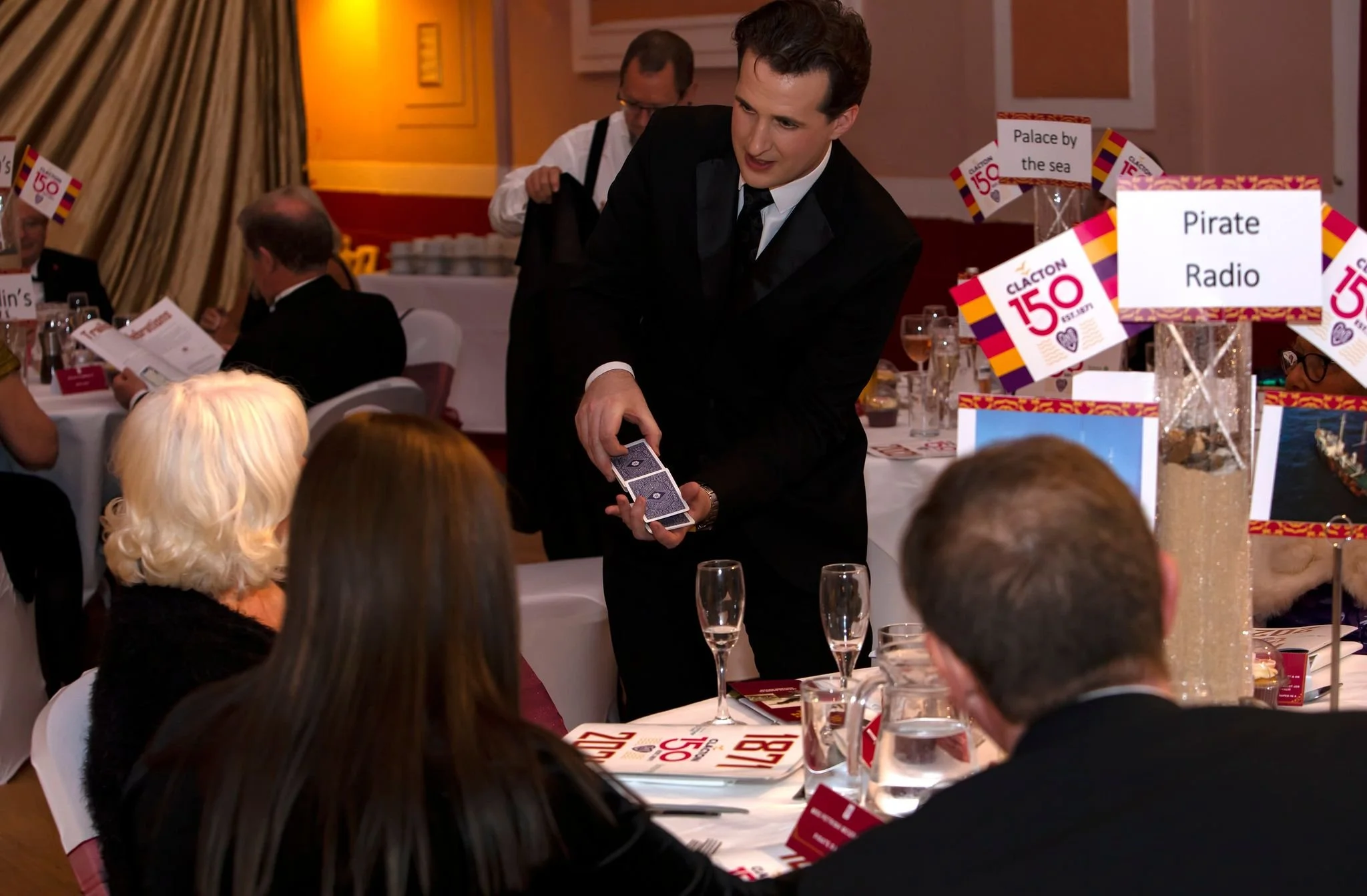 A man in a black tuxedo is performing a card trick at a formal event, with multiple guests seated at round tables around him. There are posters and signs on the tables indicating a celebration of Claction 150, and a sign that reads 'Pirate Radio.' Th