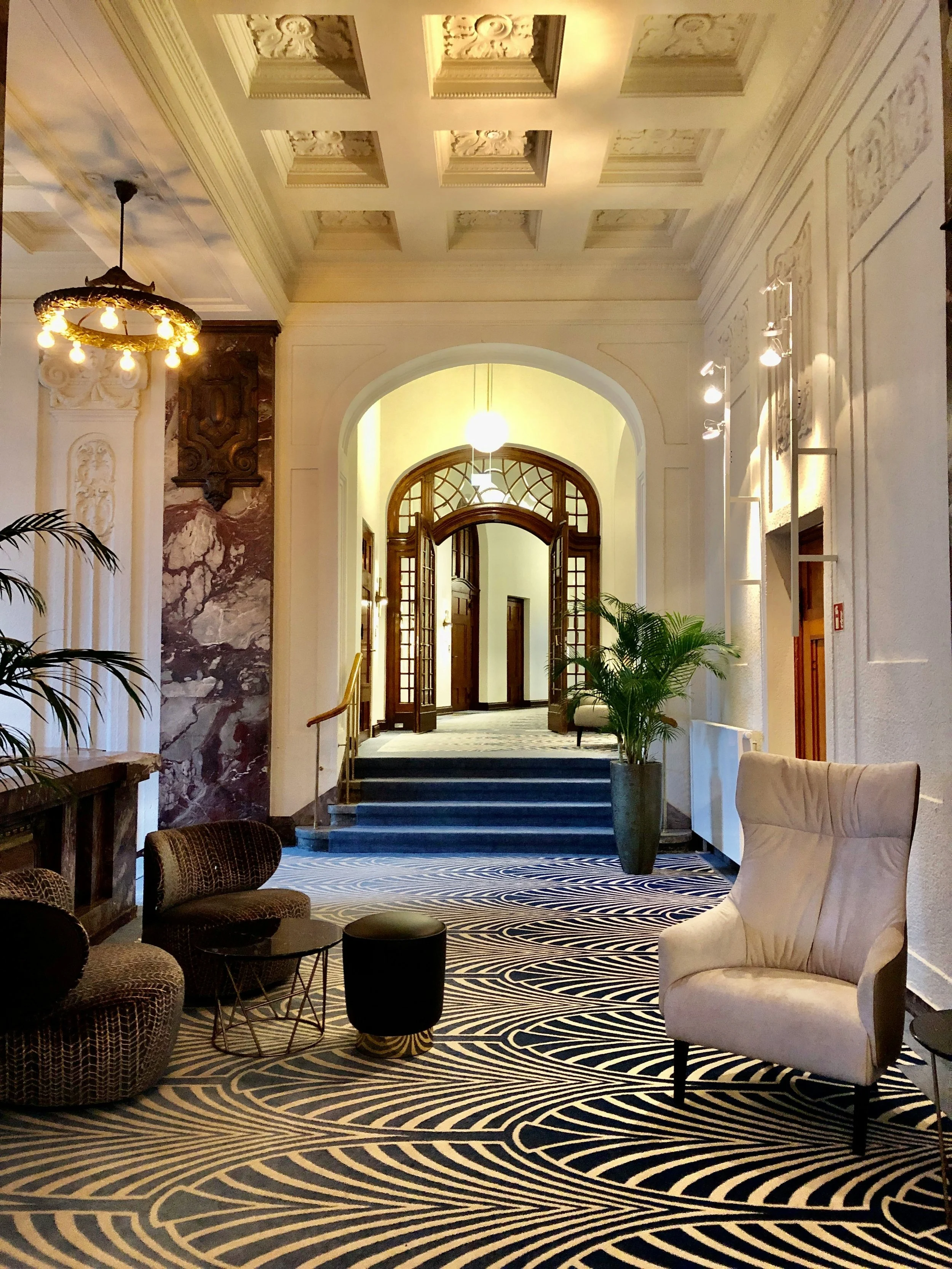L'interno di un'elegante hall di un palazzo, con soffitto decorato, lampadari, sedie imbottite e un grande vaso con piante verdi, pavimento con disegno a onde.