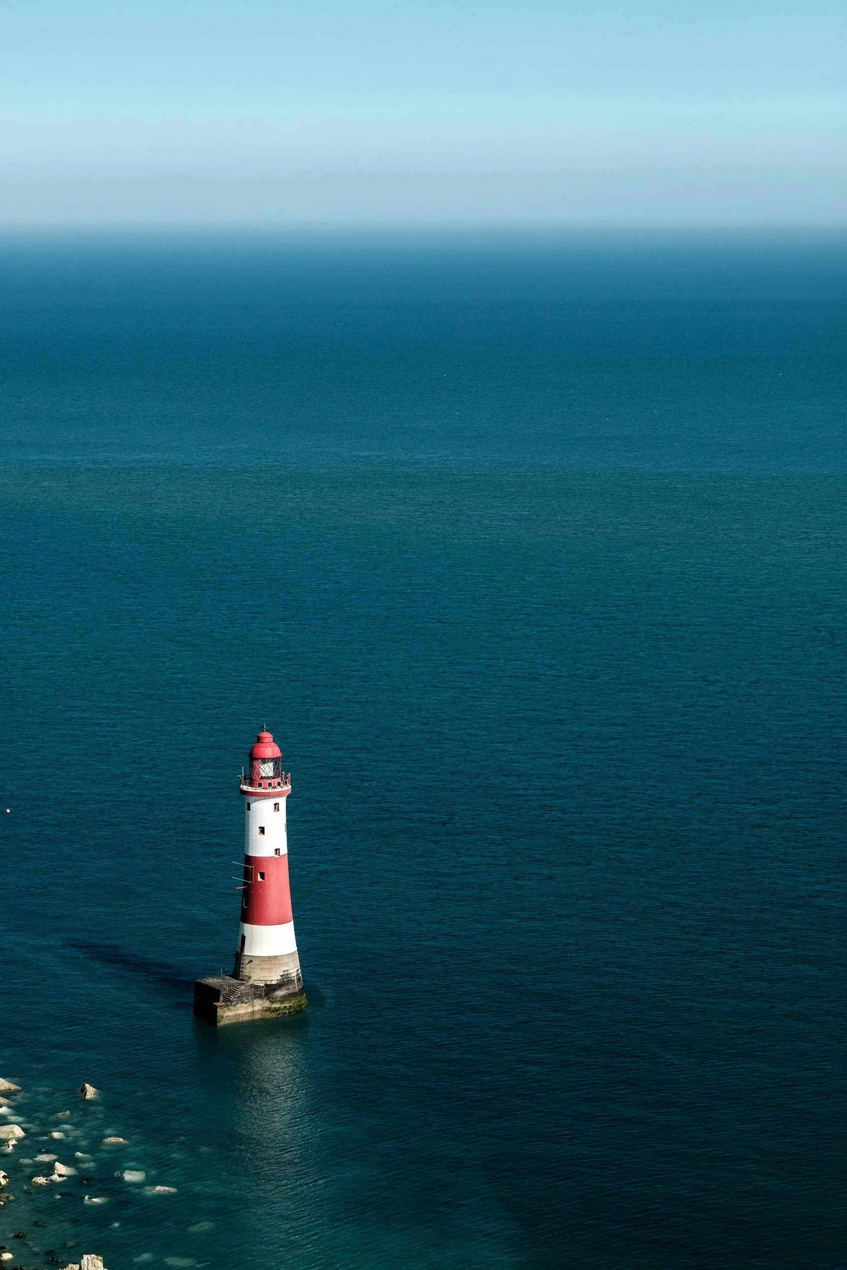 A tall lighthouse with red and white bands stands in the ocean, surrounded by water and some rocks near the shore, under a clear sky.
