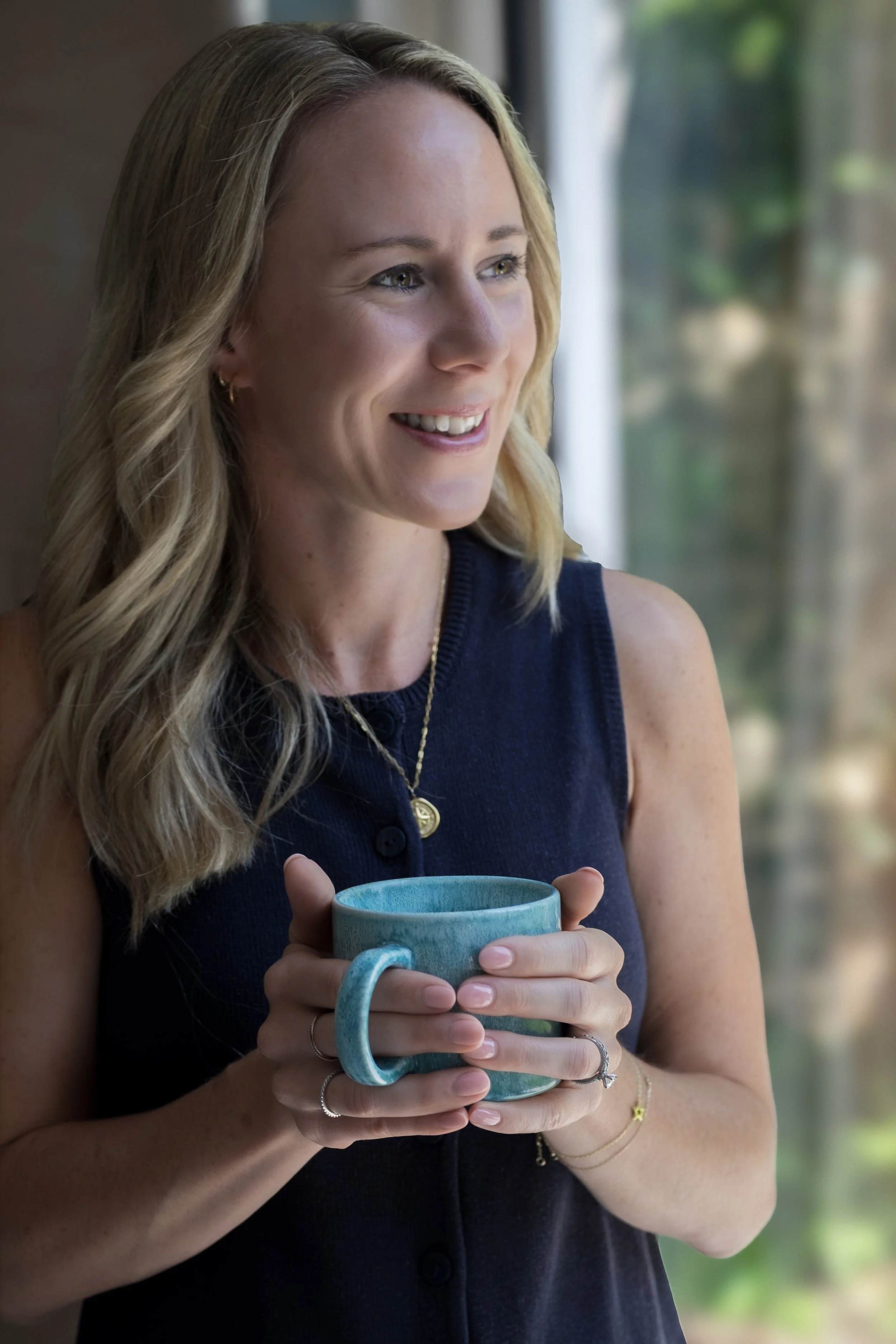 A woman with blonde hair holding a turquoise mug and smiling, standing indoors near a window.