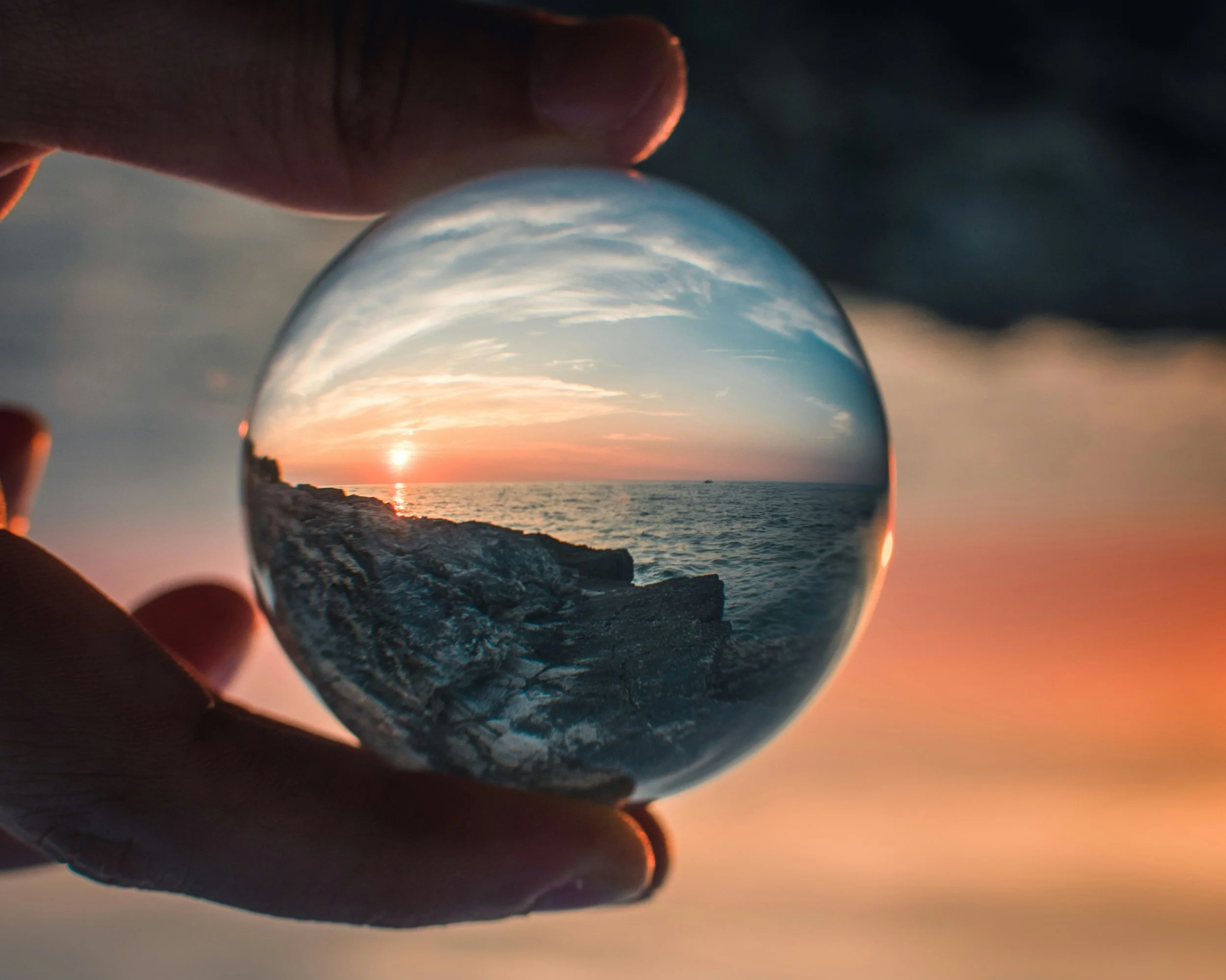 Hand holding a glass sphere reflecting a sunset over the ocean with rocks in the foreground.