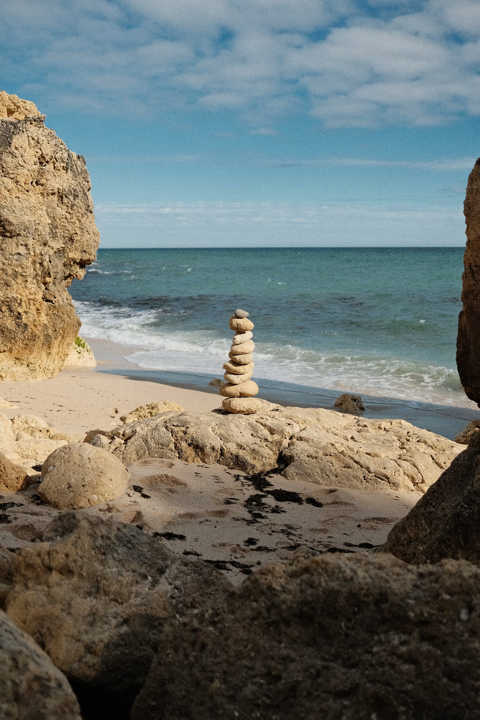Stacked stones on a sandy beach with ocean waves and a partly cloudy sky in the background.