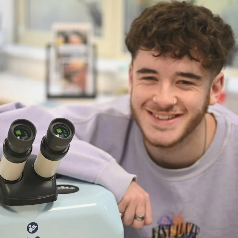 A young man with curly brown hair and a nose ring smiling at a microscope.
