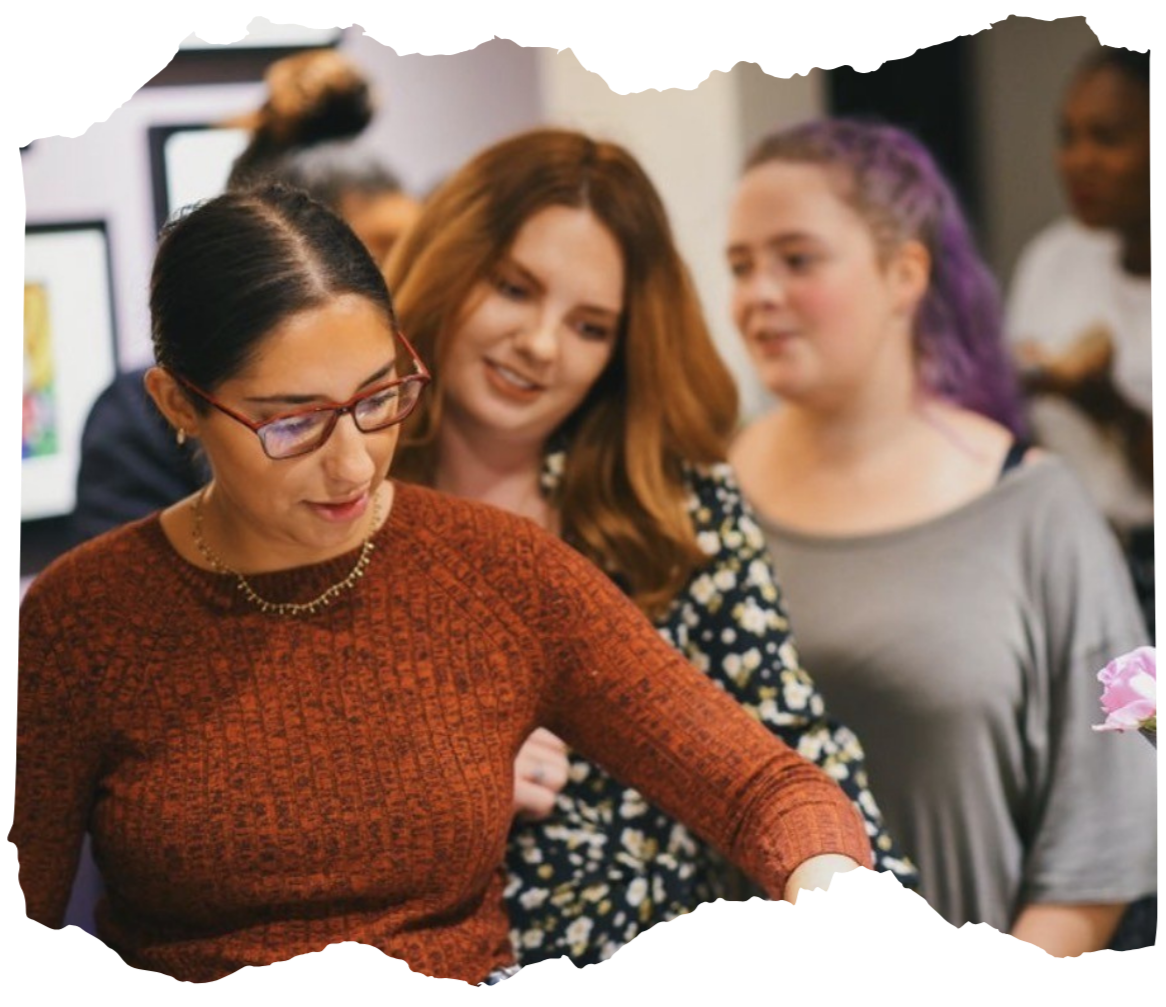 A group of women gathered around, looking at something together, in a social setting, with one woman in focus wearing glasses and a rust-colored sweater.