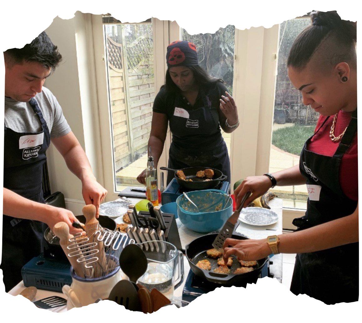 Three people cooking together in a kitchen, frying food in a skillet, with bowls and kitchen utensils on the counter, and a large window in the background showing an outdoor garden.