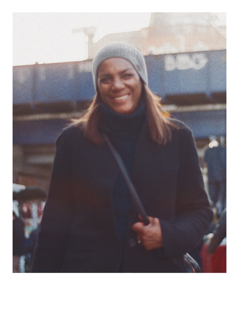 A woman wearing a gray beanie, black coat, and carrying a black bag outdoors, smiling at the camera.