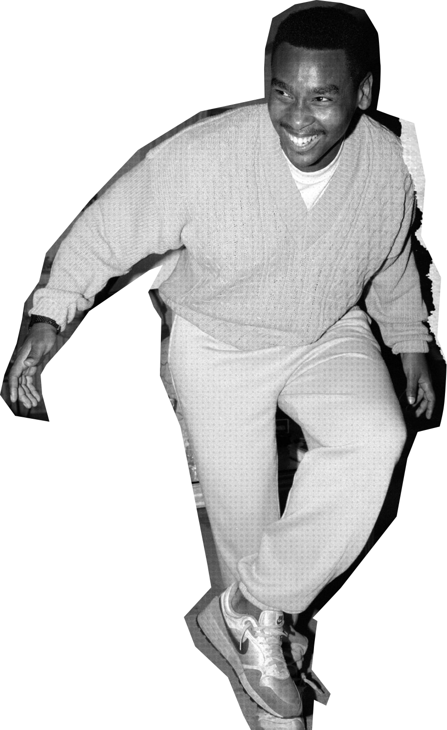 A man smiling, crouching, and leaning forward slightly, wearing a light-colored sweater, matching pants, sneakers, and a watch.