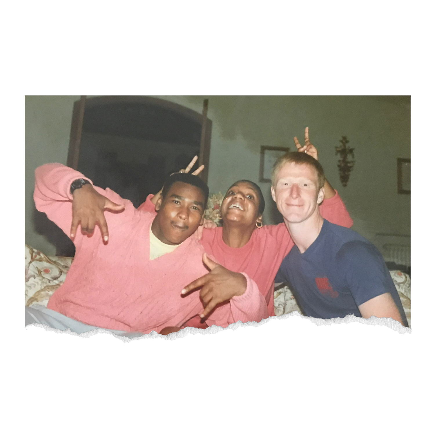 Three friends smiling and posing playfully for a photo in a cozy indoor setting, with one making peace signs and another making a rock sign.