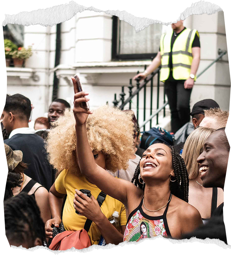 A group of people smiling and taking a selfie during an outdoor event, with a woman in the center holding her phone up. The crowd appears diverse in age and ethnicity, and a person in a reflective vest is standing on stairs in the background.
