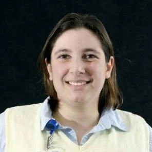 Sally, a young lady, with shoulder-length brown hair, smiling, wearing a light-coloured sweater over a collared shirt, against a dark background.
