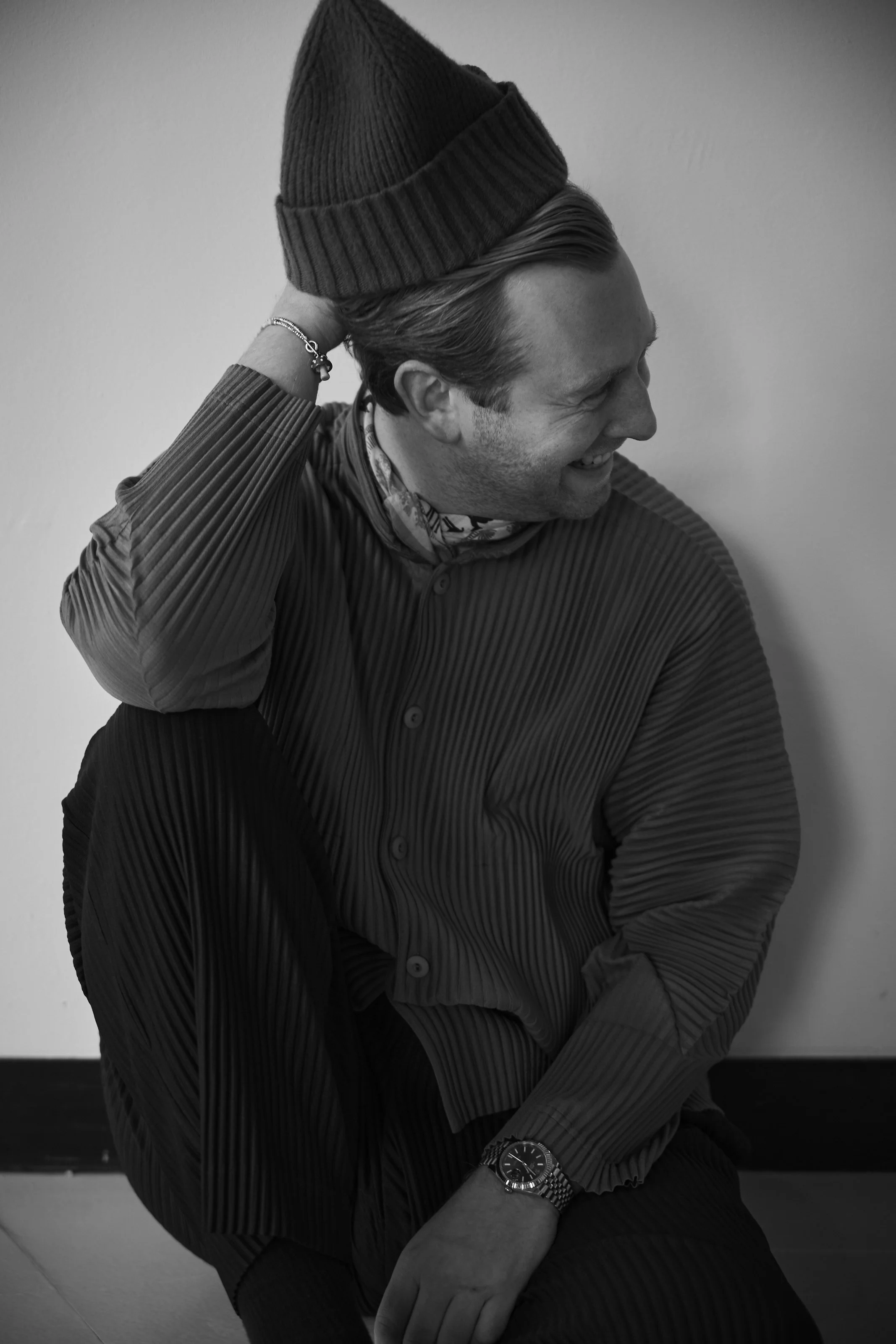 A man with slicked-back hair smiling while adjusting a beanie on his head, wearing a striped shirt and a watch on his wrist, in a black-and-white photo.