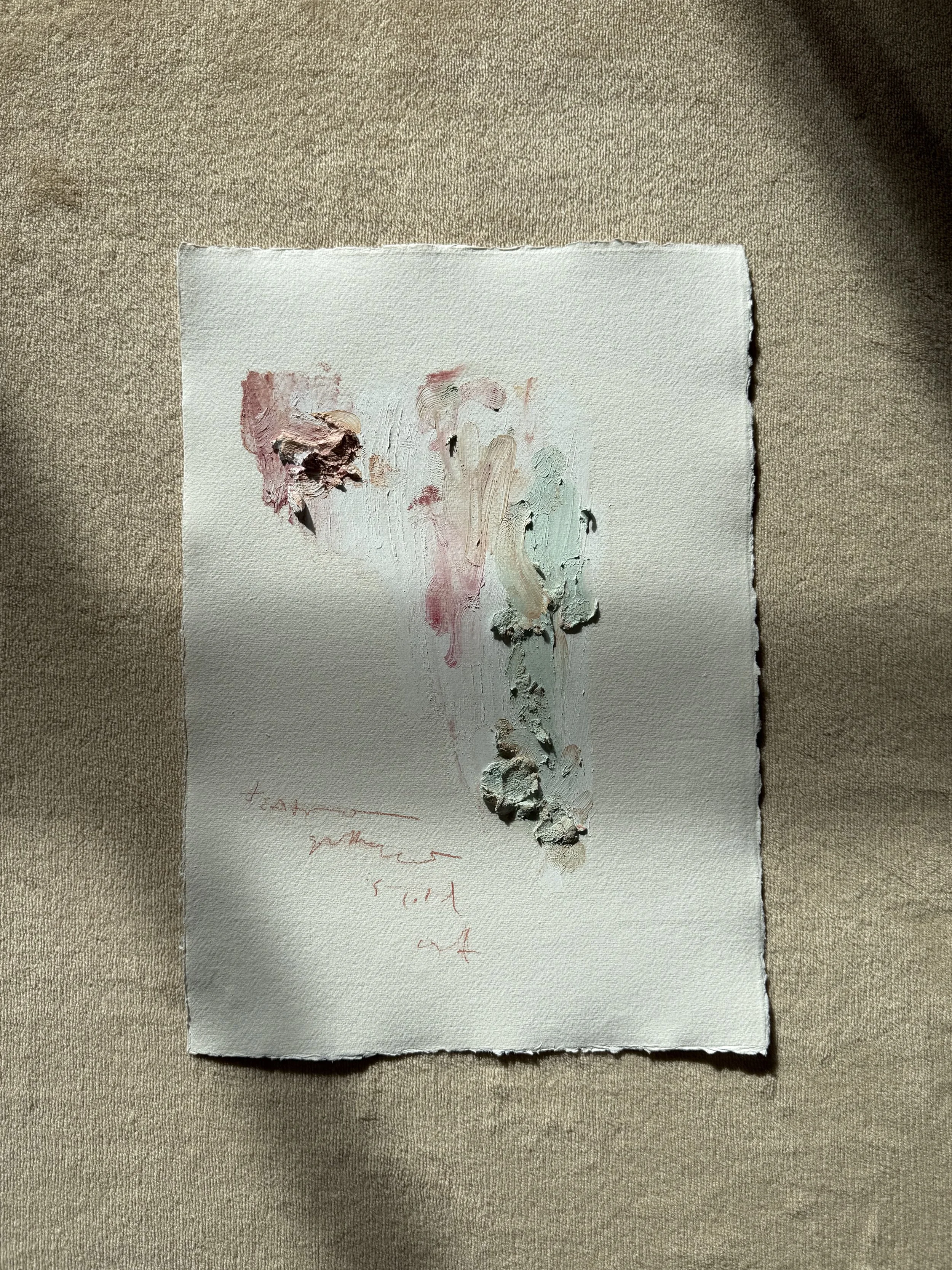 A textured piece of white paper with a small, abstract dollop of pink, beige, black, blue, and white paint in the upper left corner, placed on a beige fabric surface.