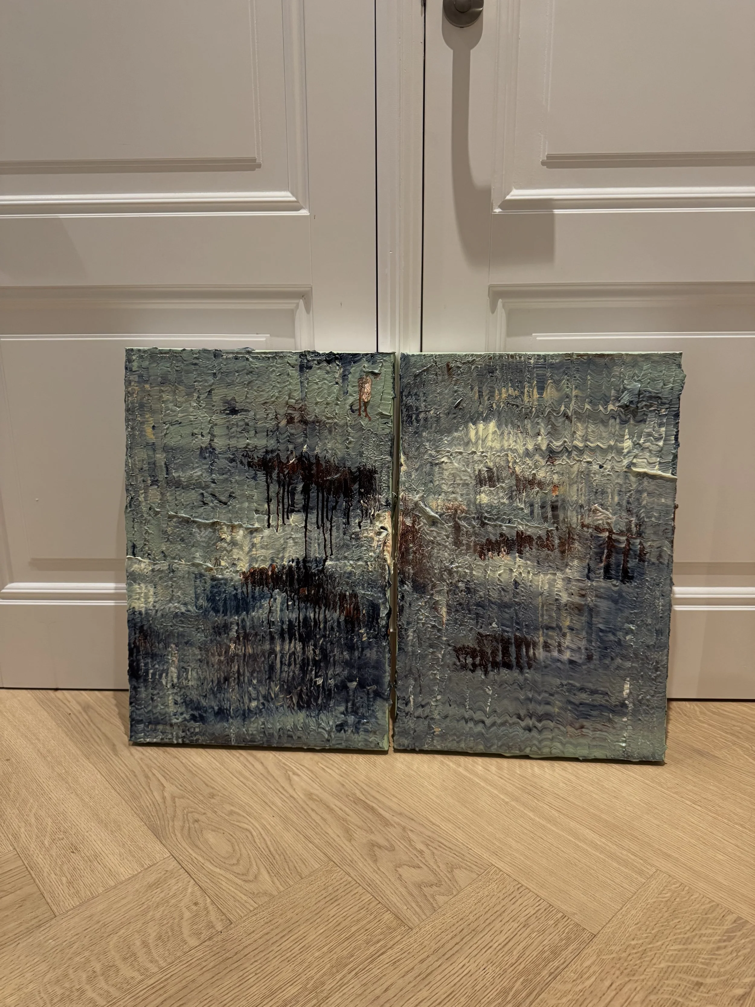 Two small abstract landscape paintings leaning against a white door with molding. The paintings have textured, thick paint with dark, muted colors, and are set on a light wood floor.