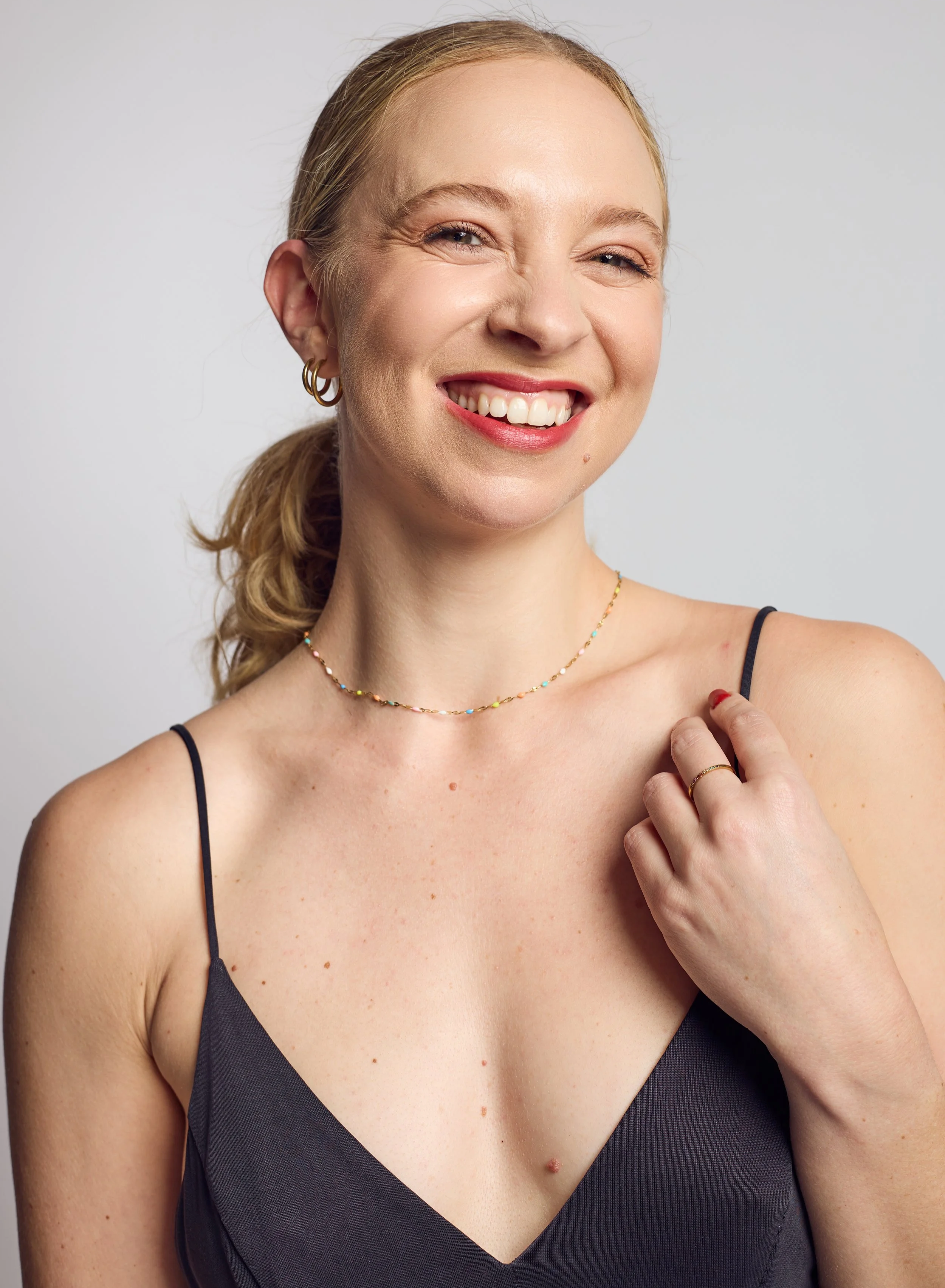 Close-up portrait of a smiling woman with light skin, blonde hair tied back, wearing a black spaghetti strap top, layered gold jewelry, and a subtle makeup look, against a plain white background.