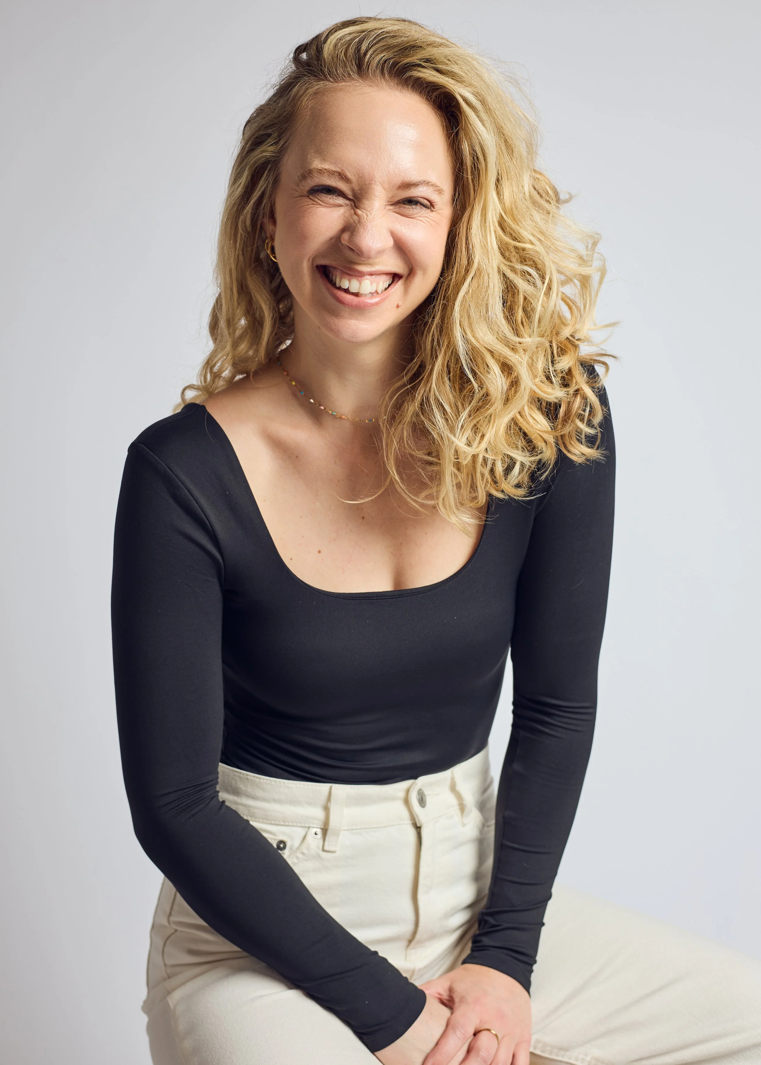 A woman with blonde curly hair wearing a black long-sleeve top and cream-colored pants, sitting against a light gray background, smiling and winking.