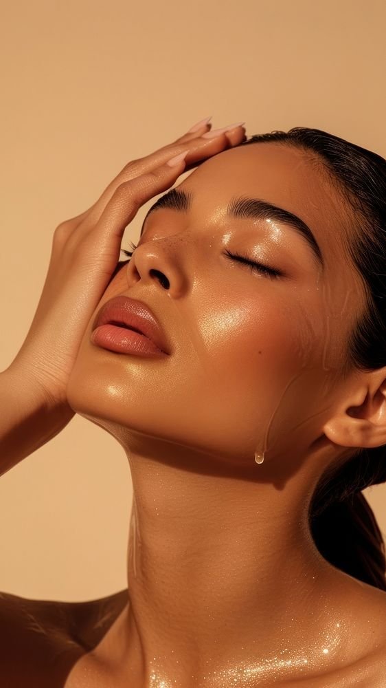 A woman with closed eyes and glowing skin touches her forehead, with water droplets on her face and neck, against a beige background.