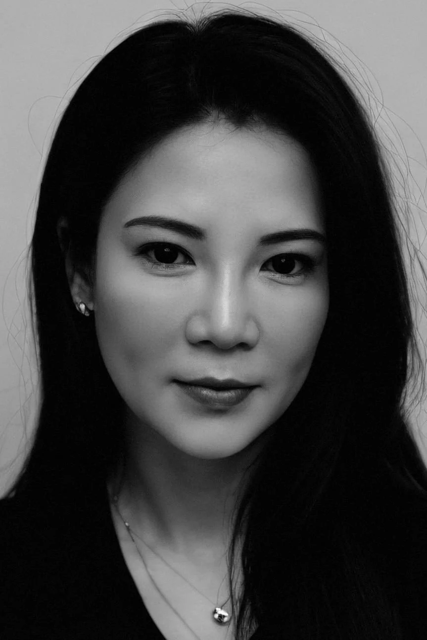 A black-and-white portrait of a woman with long dark hair, wearing earrings and a necklace, looking directly at the camera with a slight smile.