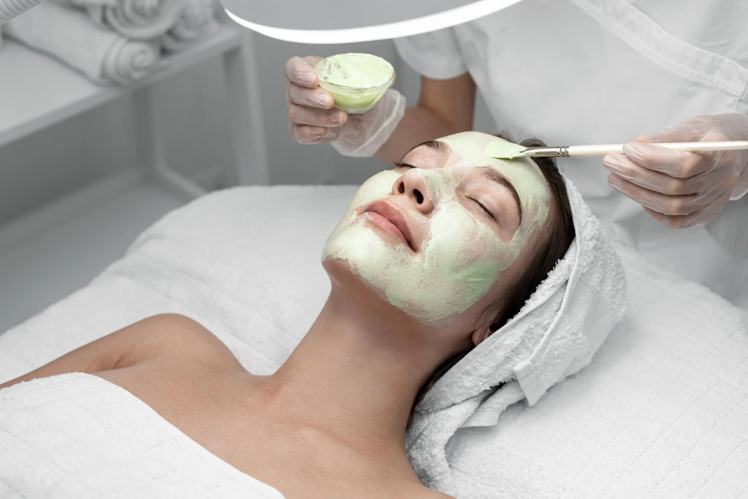 A woman lying on a spa bed with eyes closed, getting a facial treatment. A person in white attire is applying a green facial mask with a brush and holding a bowl of the mask.