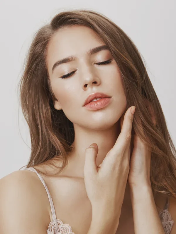 A woman with long brown hair is posing with her eyes closed, one hand gently touching her face.