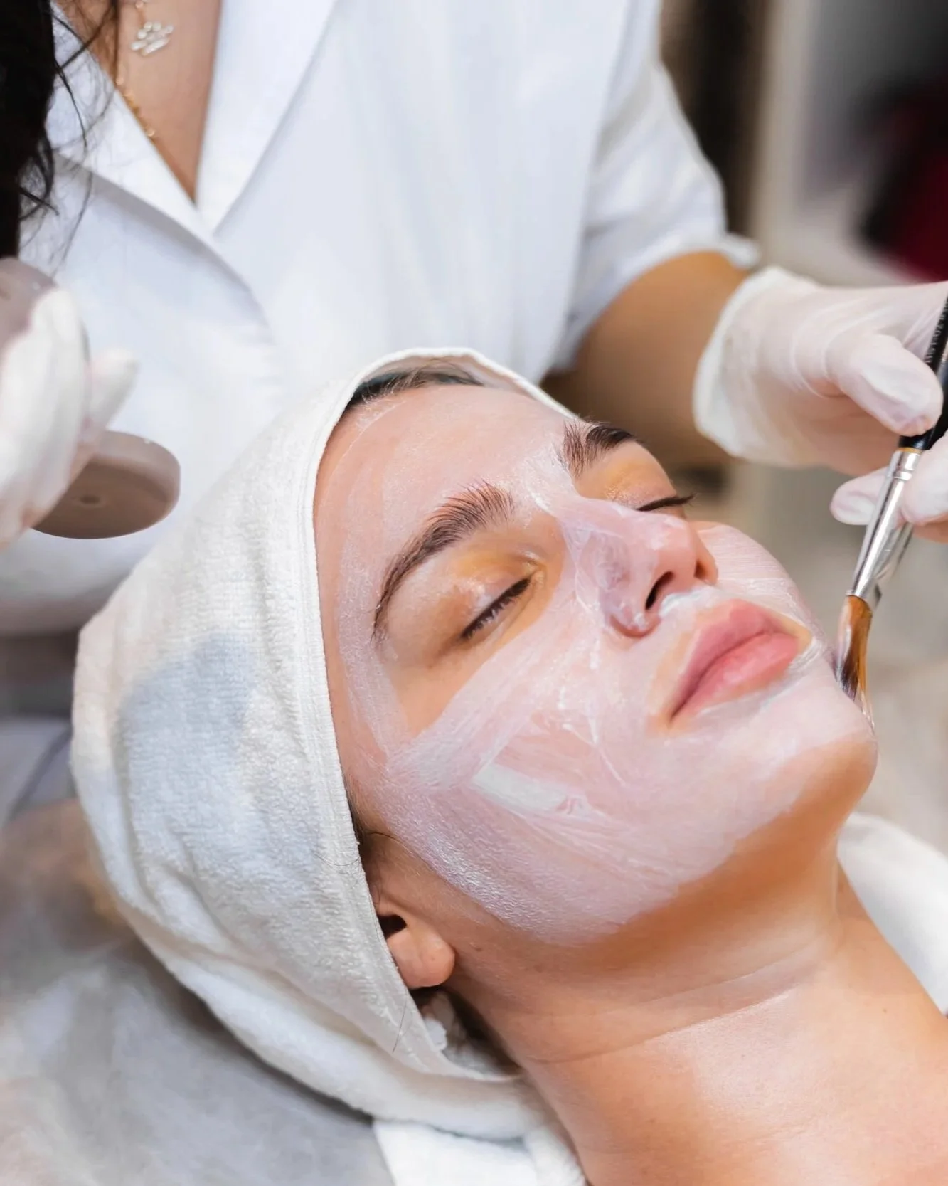 A woman with a white towel wrapped around her head and face, lying down with closed eyes while a professional applies a skincare treatment using a brush.