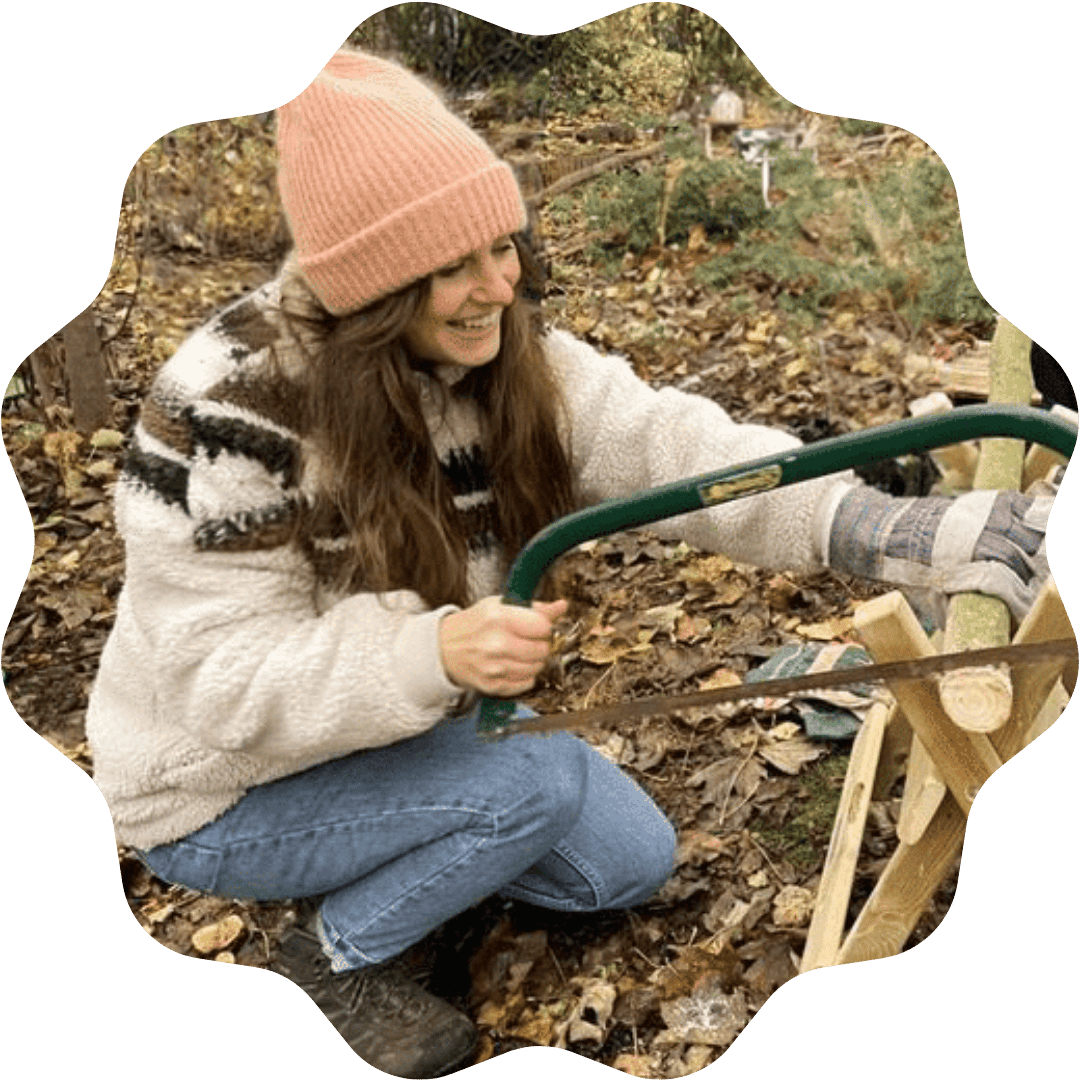 A woman wearing a pink knit hat, plaid shirt, and jeans, using a saw to cut wood outdoors among fallen autumn leaves.
