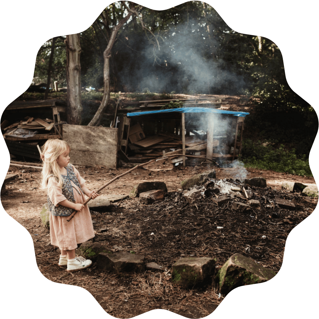 A young girl with blonde hair in a pink dress and white shoes, holding a stick, stands near a small smoky fire outdoors in a wooded area with a wooden structure and trees in the background.