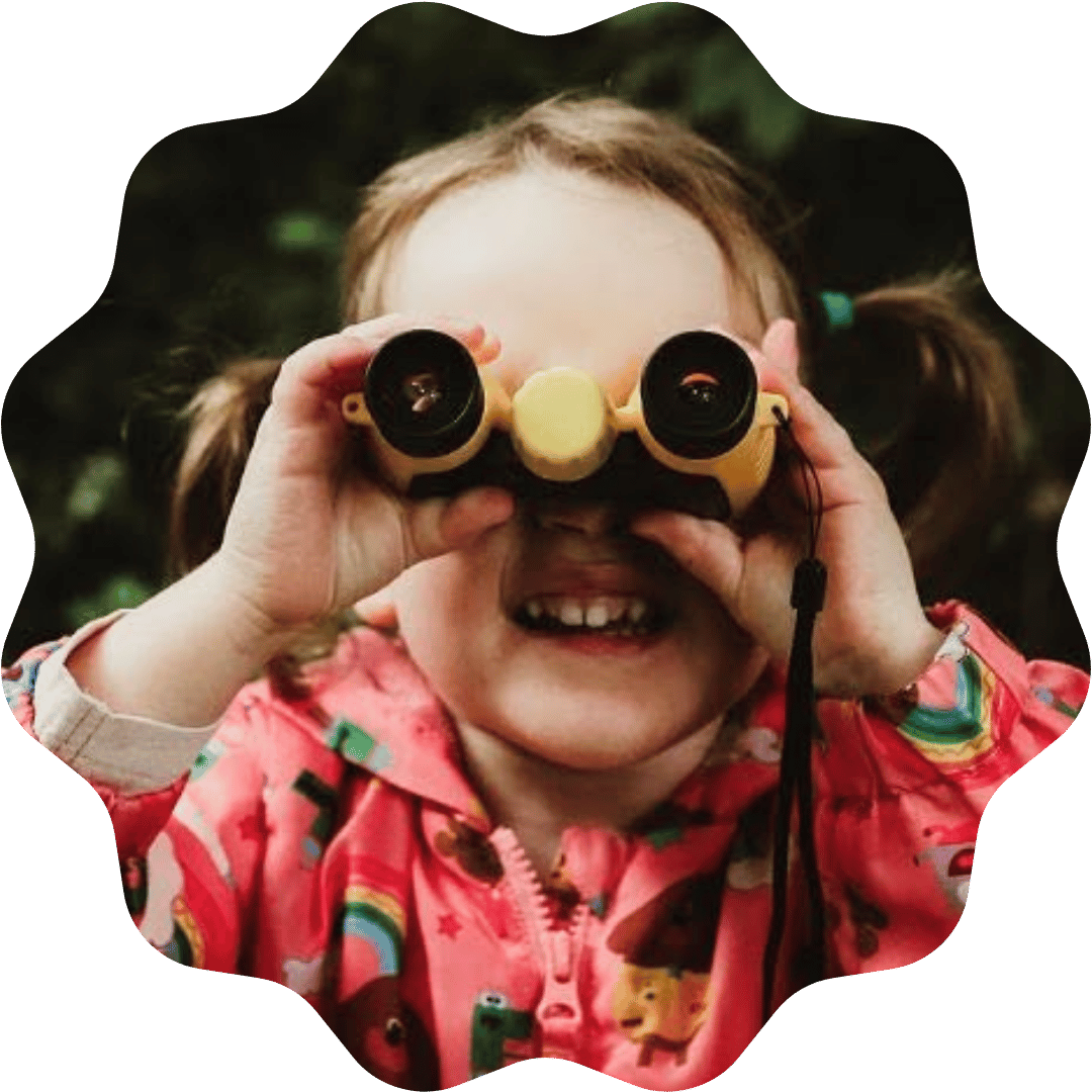 A young girl with pigtails, wearing a pink jacket with colorful patterns, looking through yellow binoculars with black lenses and smiling outdoors.