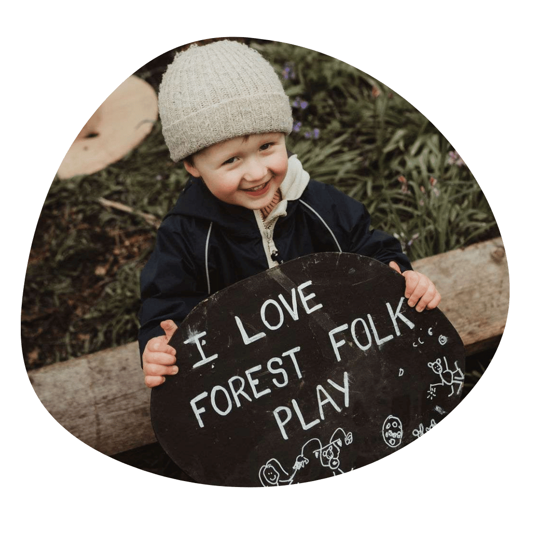 A young child smiling and holding a black sign that says 'I love forest folk play,' outdoors with greenery and flowers in the background.