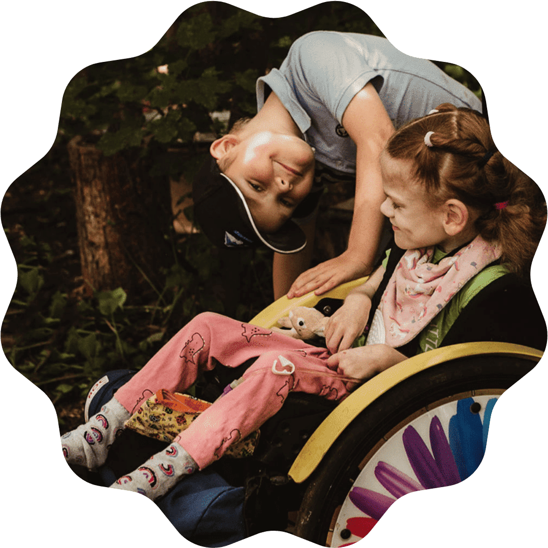 A young boy with a hat leaning over a girl in a wheelchair, smiling and touching her arm outdoors among trees.