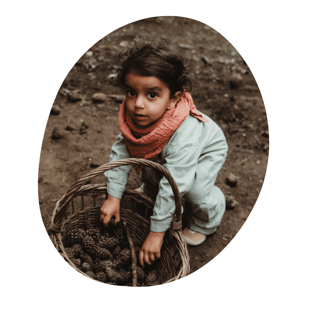 A young child with curly dark hair, wearing a pink scarf and light-colored jacket, crouches down outdoors next to a basket filled with pinecones, looking at the camera.