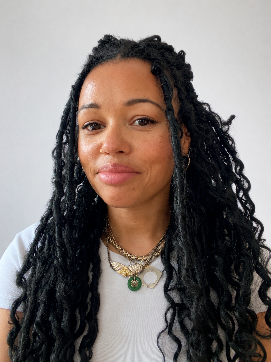 A woman with long, curly black hair wearing layered necklaces, a white top, and small hoop earrings against a plain background.