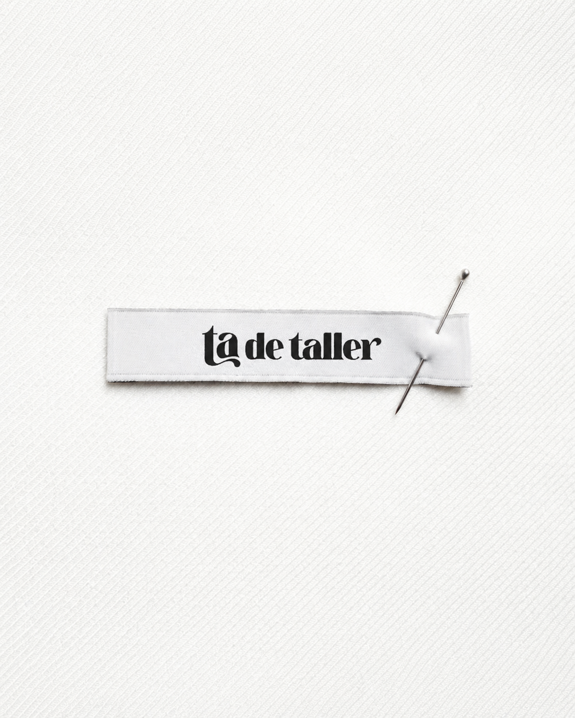 Fabric label with the text "la de taller" pinned onto a white surface with two sewing pins.