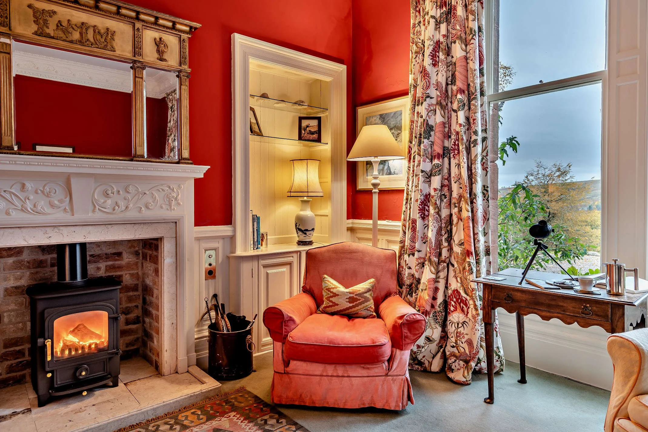 A cozy living room corner with a red wall, a fireplace with a lit stove, a patterned rug, a pink armchair with a decorative cushion, a floor lamp, a built-in shelf with framed photos, floral curtains, and a window showing trees outside.
