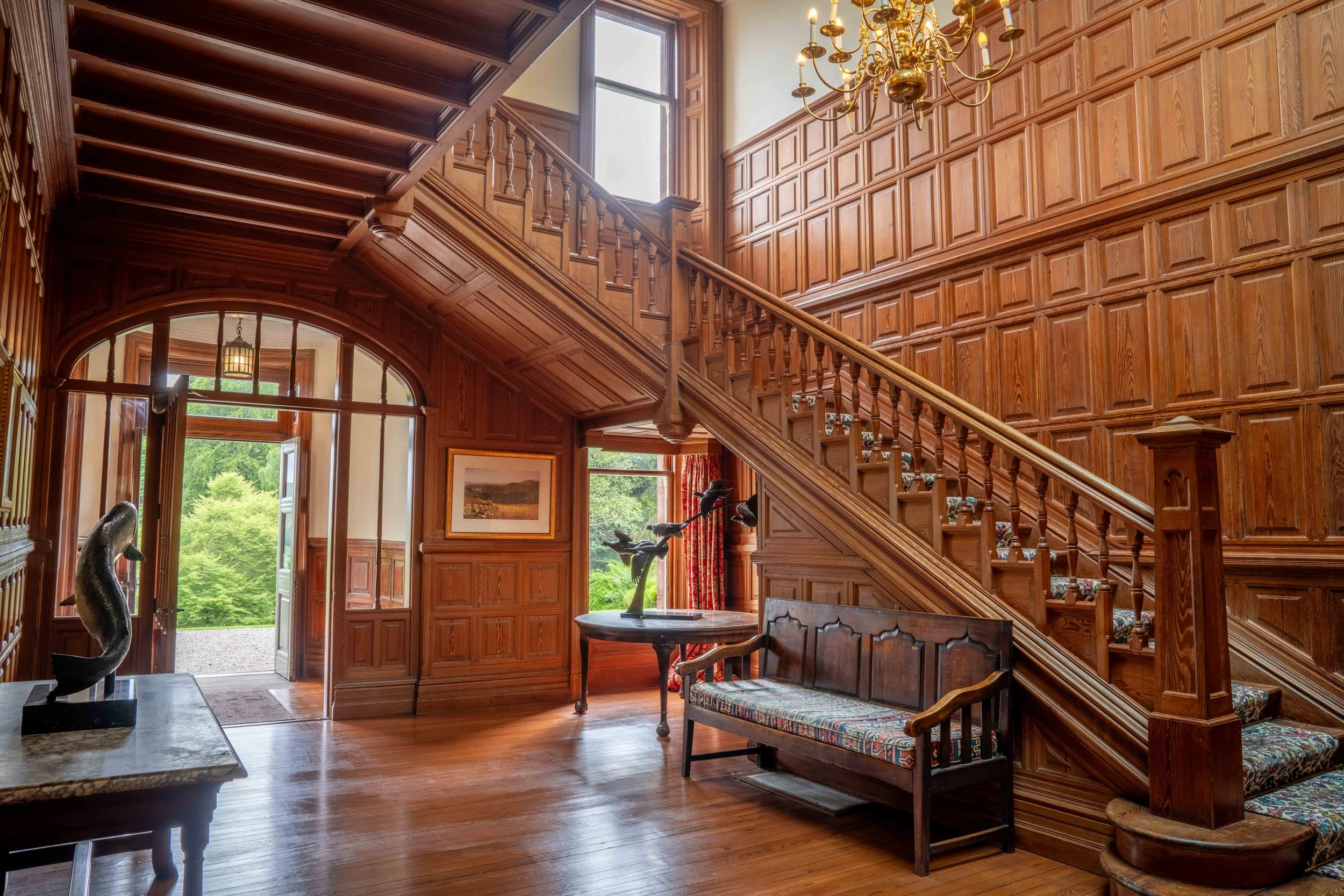 Interior of a grand wooden house with detailed wood paneling, staircase, chandelier, large window, and antique furniture.