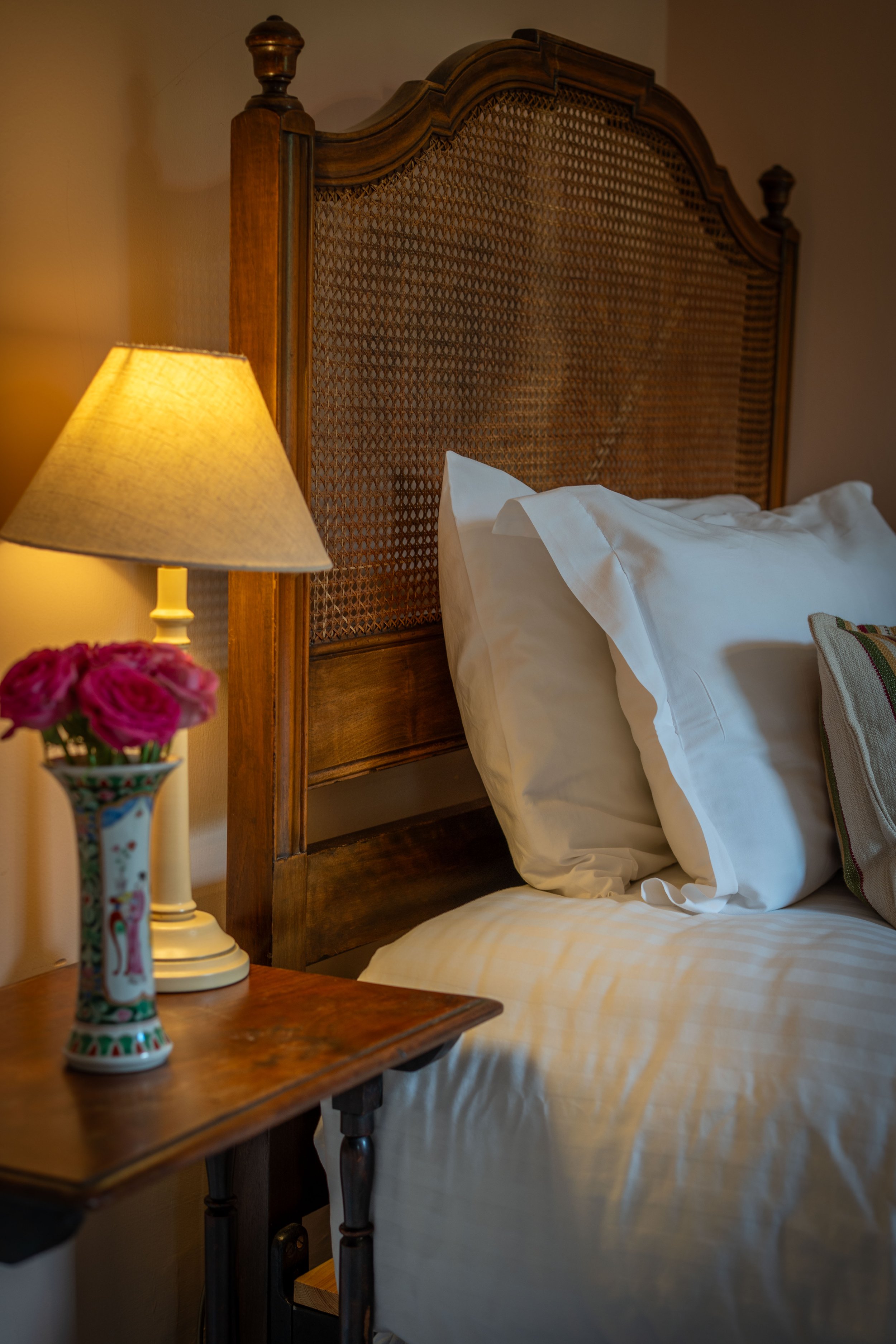 A cozy bedroom with a wooden bed featuring a woven headboard, white linens, a bedside table with a colorful flower vase, a lamp with a beige shade, and a decorative pillow.