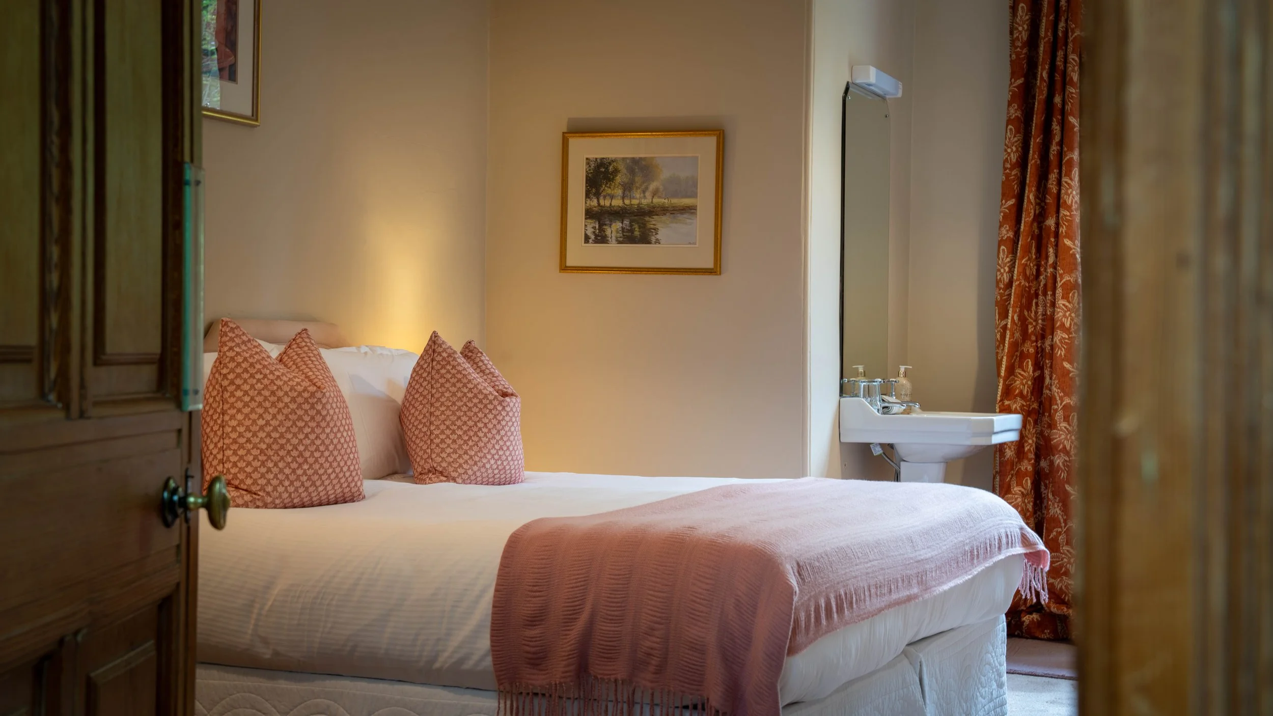 A cozy bedroom with a bed that has pink patterned pillows, a pink blanket, and a framed landscape painting on the wall. There is a small sink with soap and a mirror near the window with orange floral curtains.