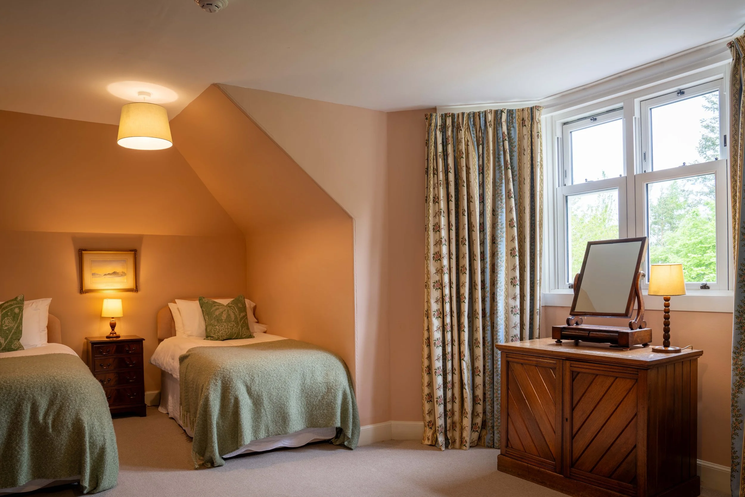 A cozy bedroom with three beds, each with green bedspreads and patterned pillows, a nightstand with a lamp, artwork on the wall, a window with floral curtains, and a wooden dresser with a mirror and a lamp.