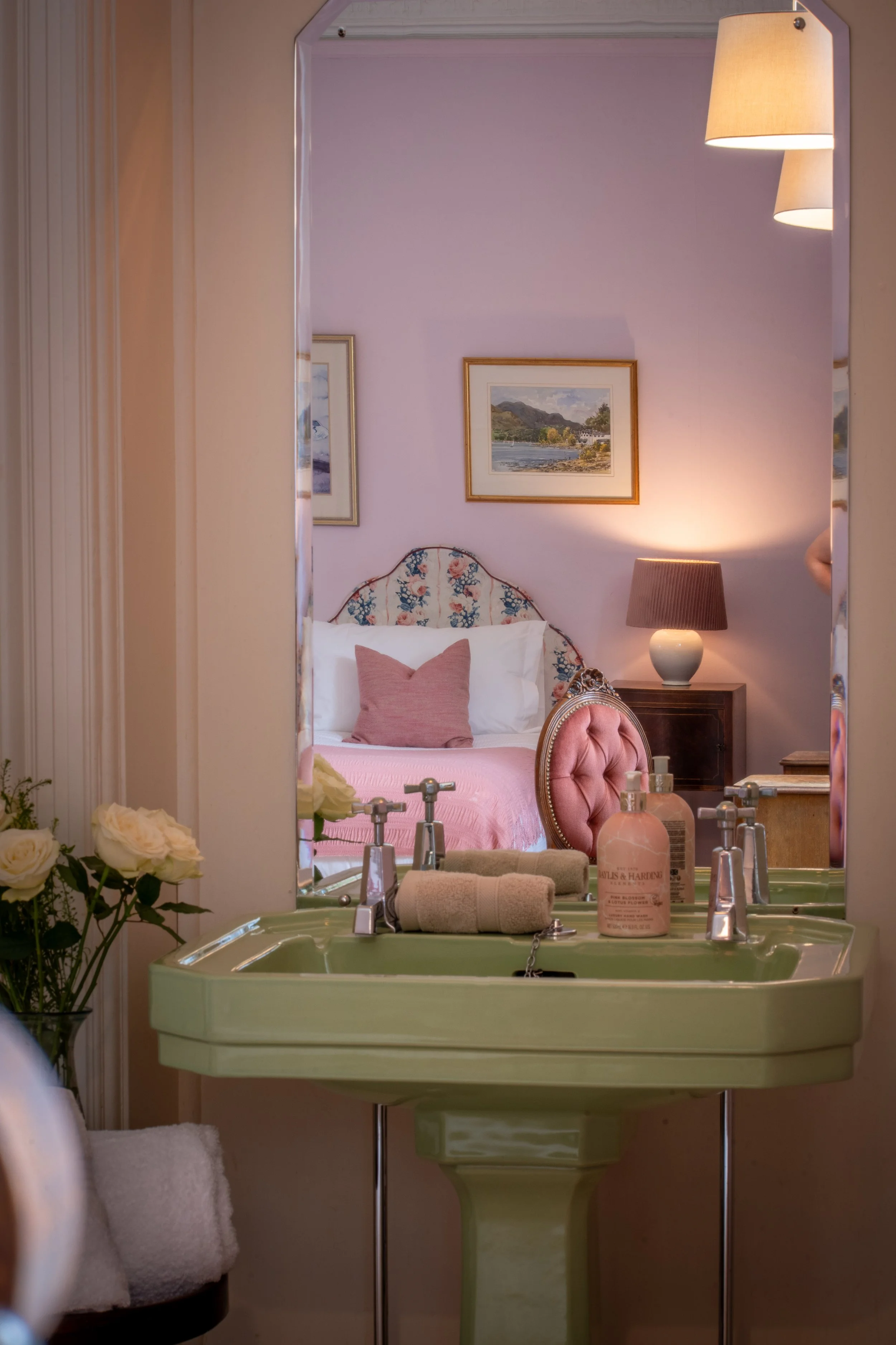A pastel green vintage sink with a floral upholstered headboard in a bedroom reflected in a mirror. The sink has soap bottles, a rolled towel, and nearby flowers. The bedroom has a pink wall, framed landscape paintings, a table lamp, and pink pillows