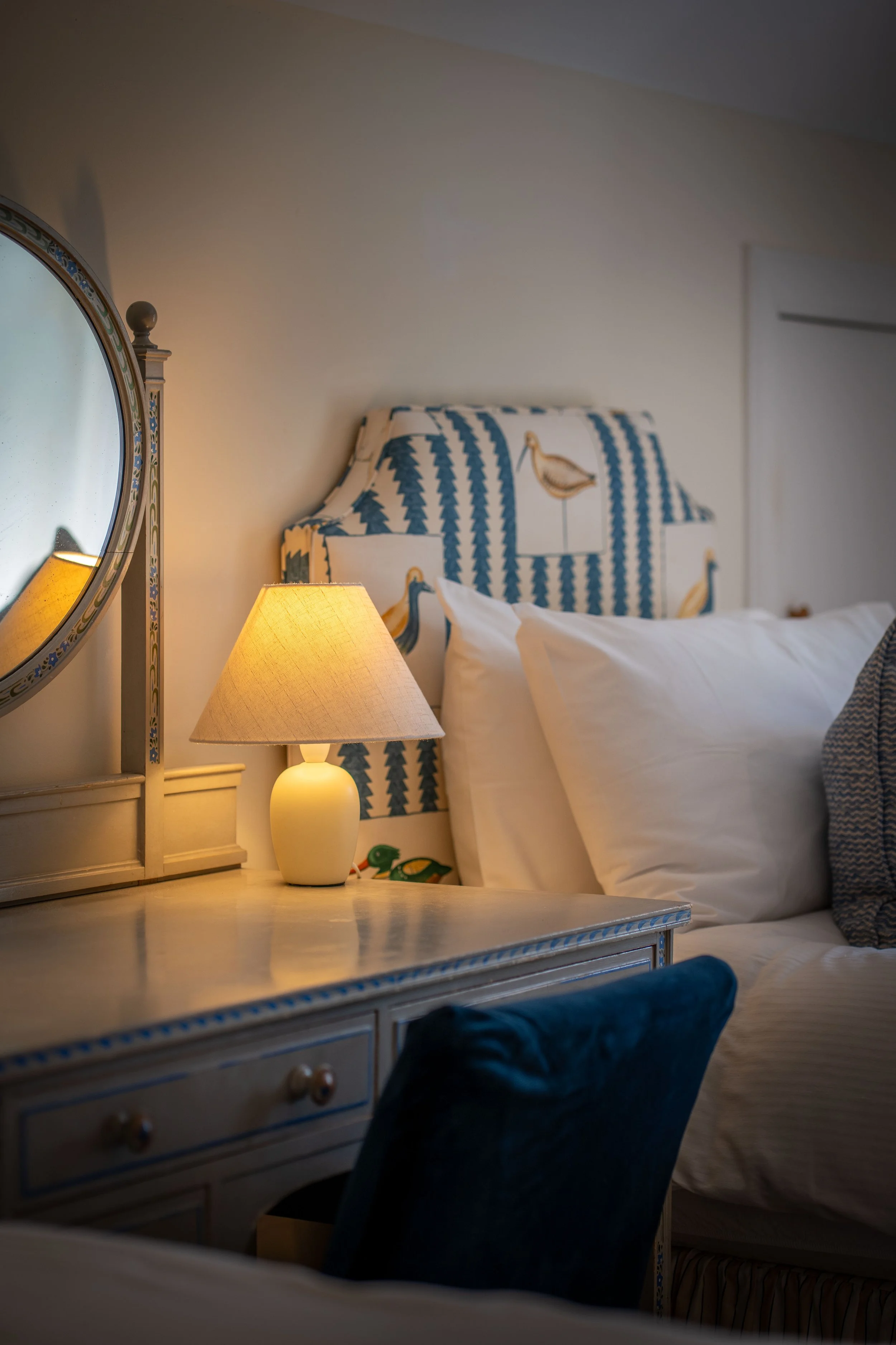 A cozy bedroom corner featuring a white nightstand with a yellow table lamp, a mirror with ornate trim, and a bed with a striped headboard decorated with bird illustrations. There is a dark blue chair in front of the nightstand.