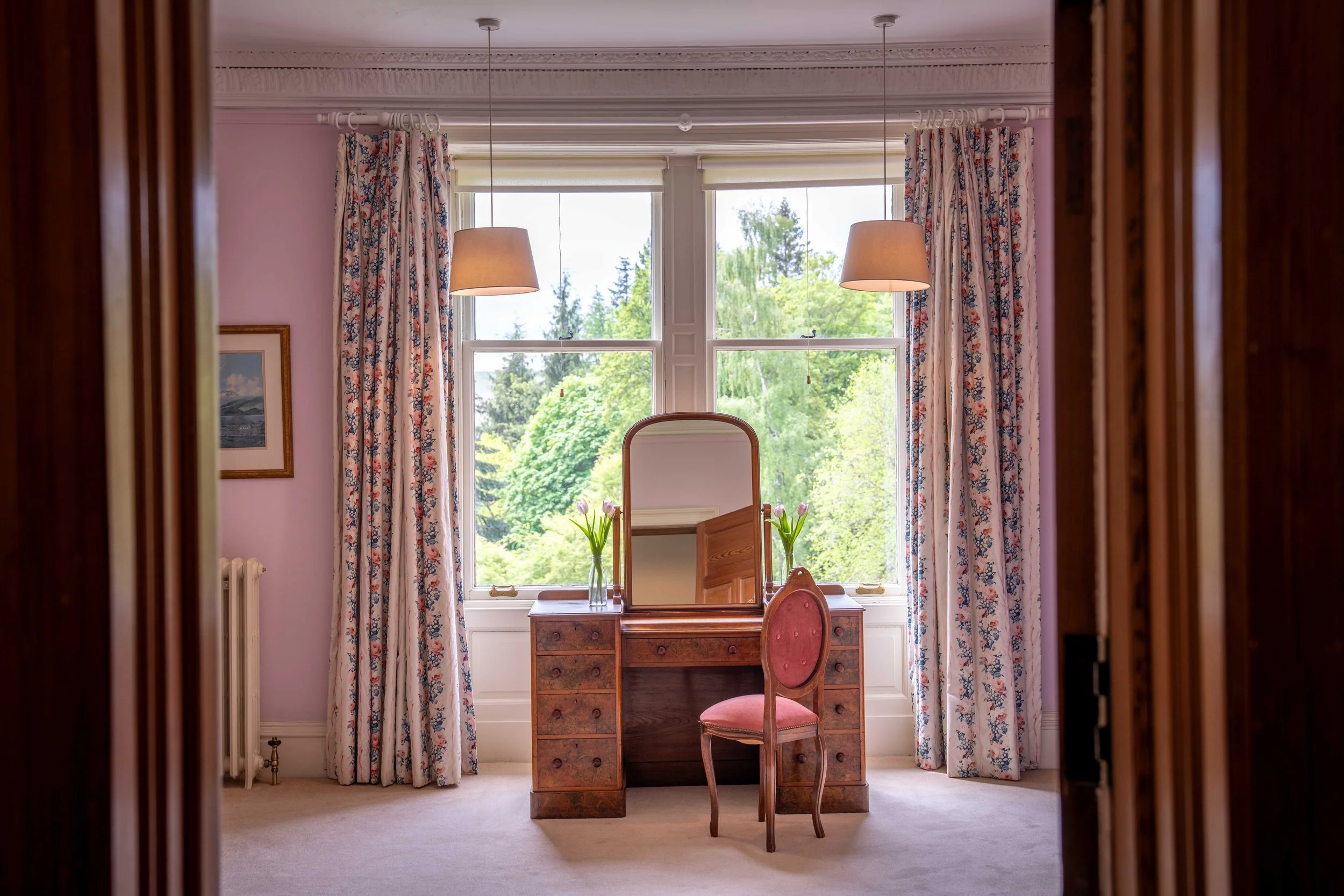 View of a room through a doorway showing a window with floral curtains, a small wooden desk with a mirror, two vases with tulips, and a pink upholstered chair. The scene outside the window includes trees and greenery.