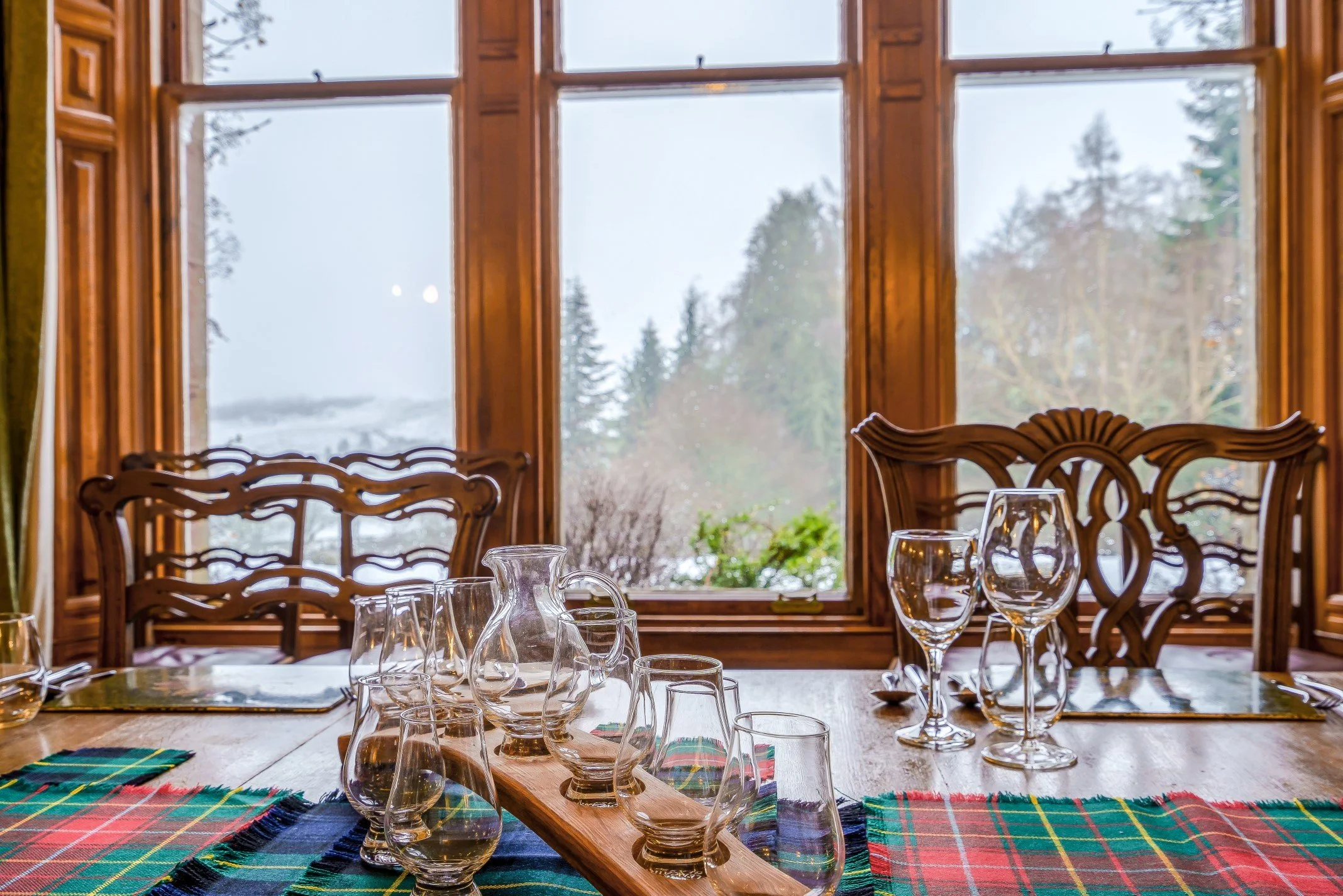 A dining table set with glassware, placed near large wooden-framed windows showing a winter landscape outside.