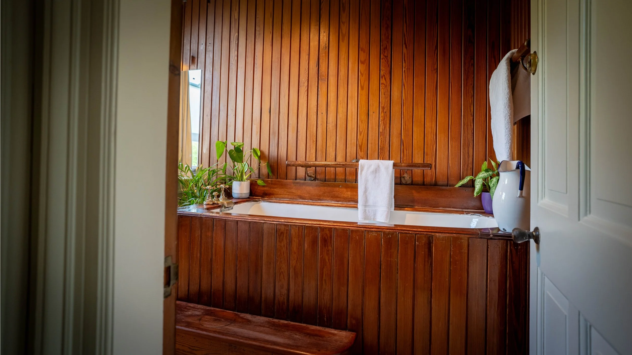 A bathroom with wooden paneling on the wall surrounding a bathtub. The bathtub has a white towel hanging on a wooden rail, and is decorated with potted plants on the edge. There is a shower curtain partially visible on the right side.